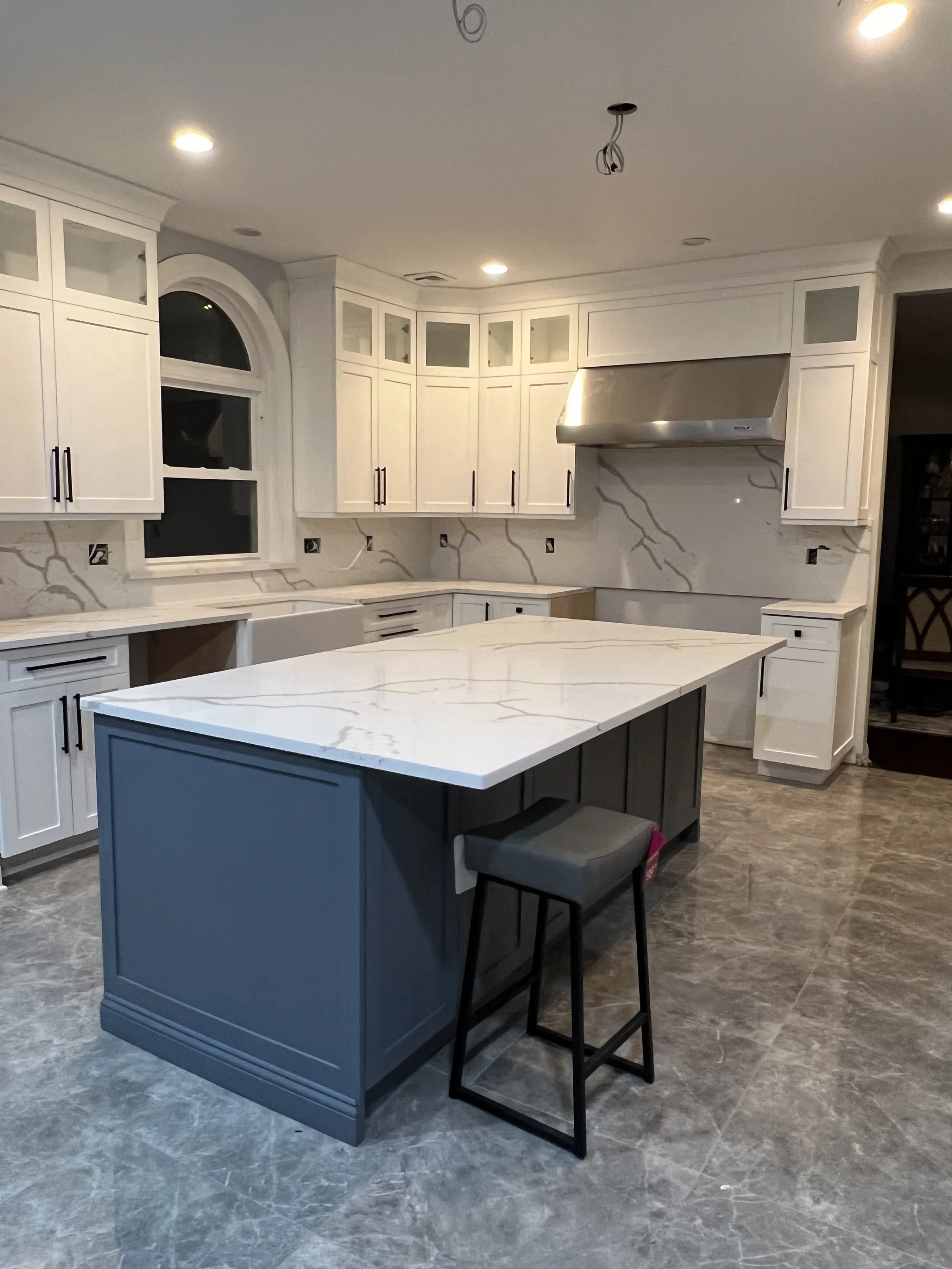 Modern kitchen with white cabinets, marble backsplash, grey island with marble countertop, black bar stool, stainless steel range hood, and recessed ceiling lights.