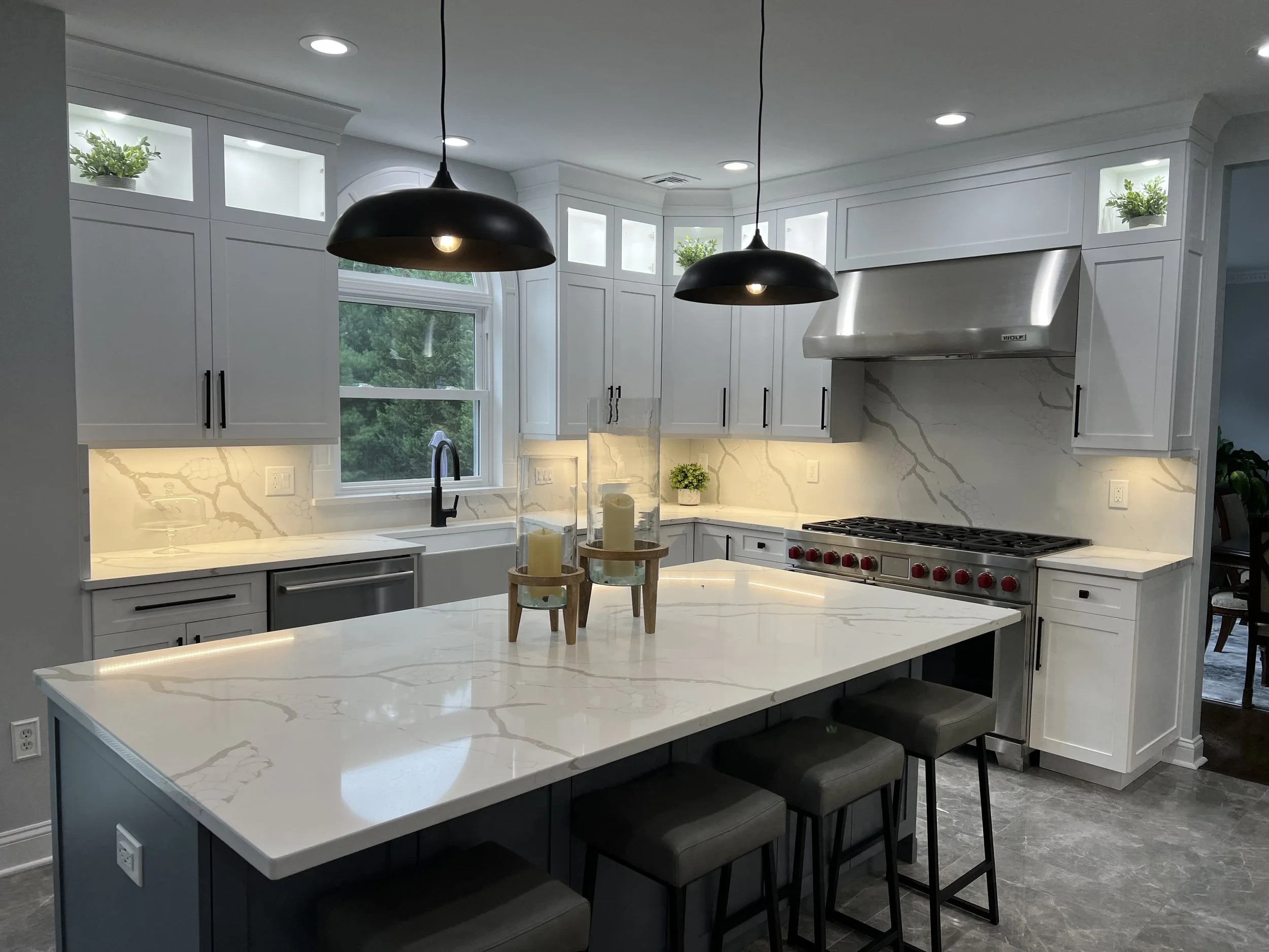 Modern white kitchen with marble countertops and backsplash, black kitchen fixtures, and black pendant lights over an island with four black stools, a large window, and potted plants.