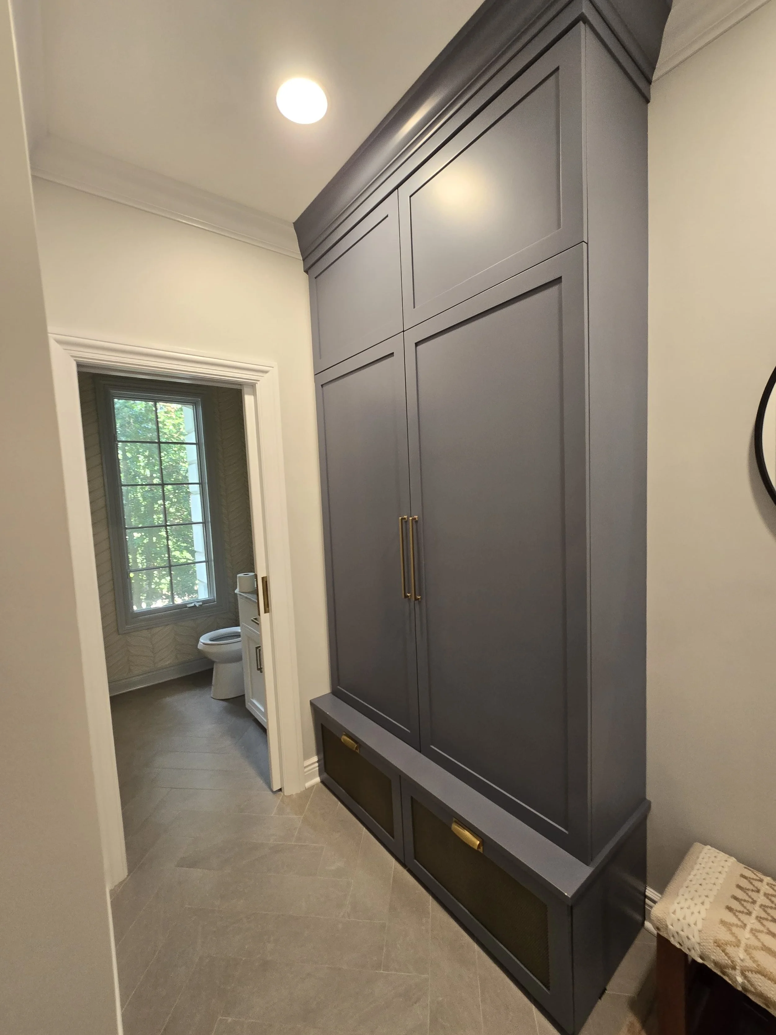 A tall gray cabinet with gold handles in a hallway, with a bathroom visible through an open door in the background, and a window with trees outside.