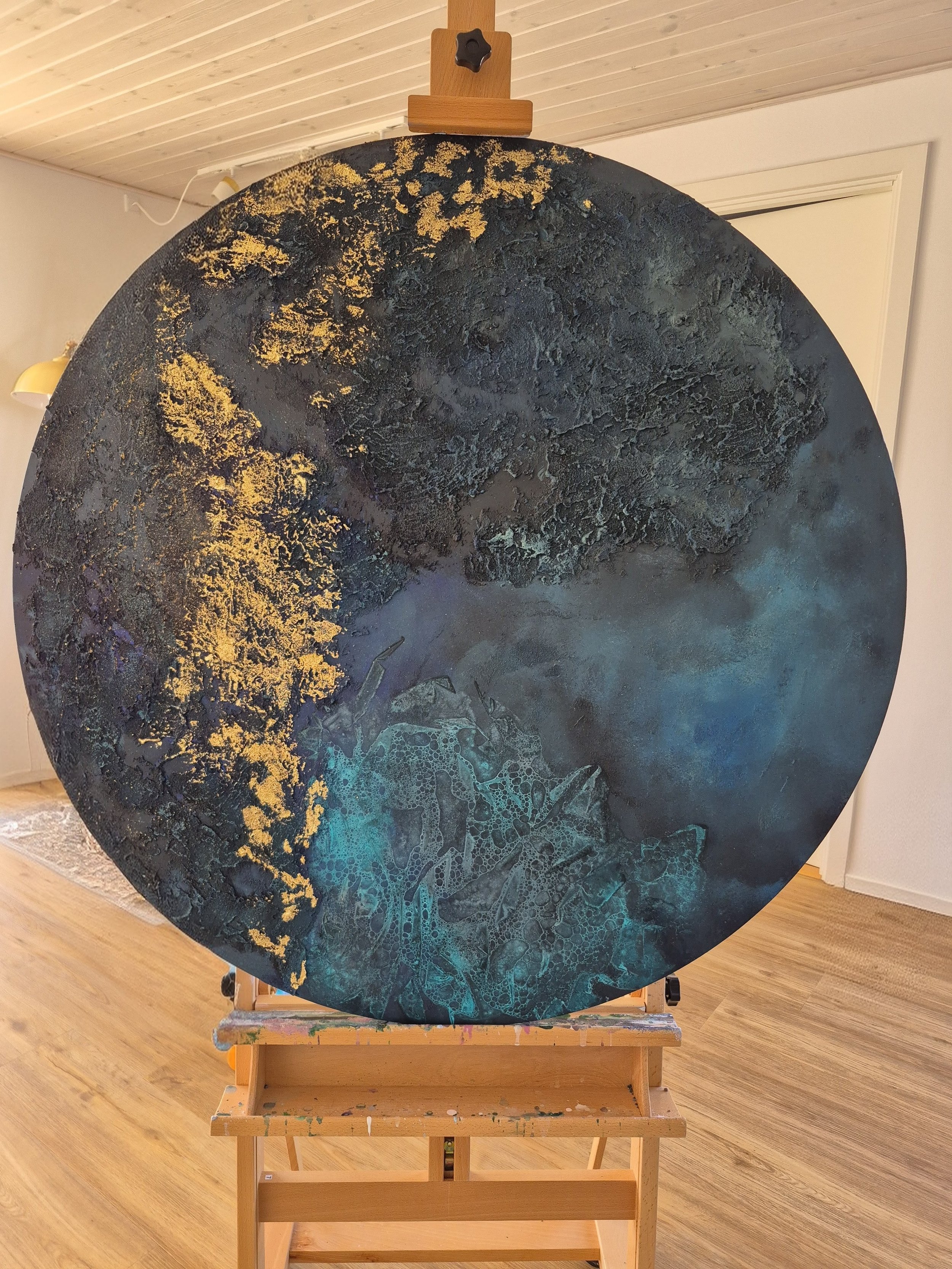 ​Modern living room featuring a large circular abstract wall art piece with deep teal, navy, and gold leaf textures hanging on a vertical wood-paneled wall above a cream-colored minimalist sofa.