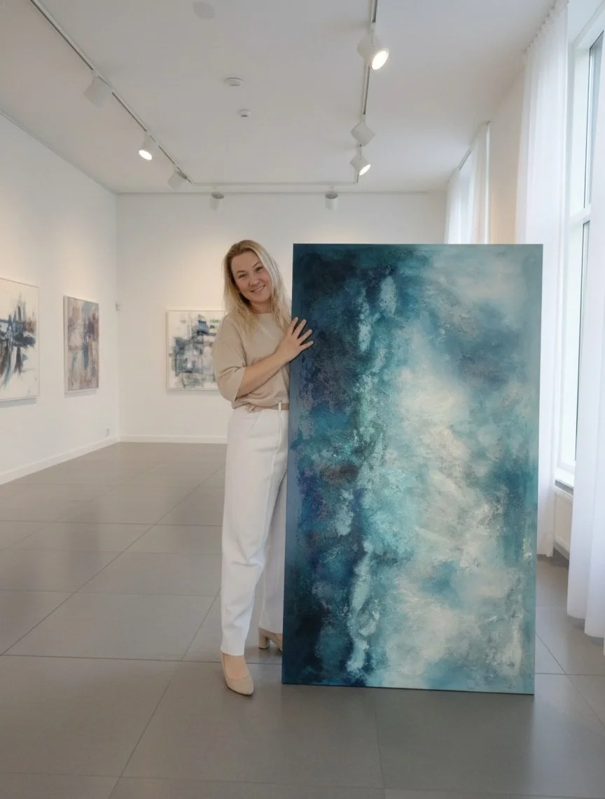 Woman standing in a bright gallery space, smiling and holding a large abstract painting in shades of blue, teal, and white. Other abstract artworks are visible on the walls in the background