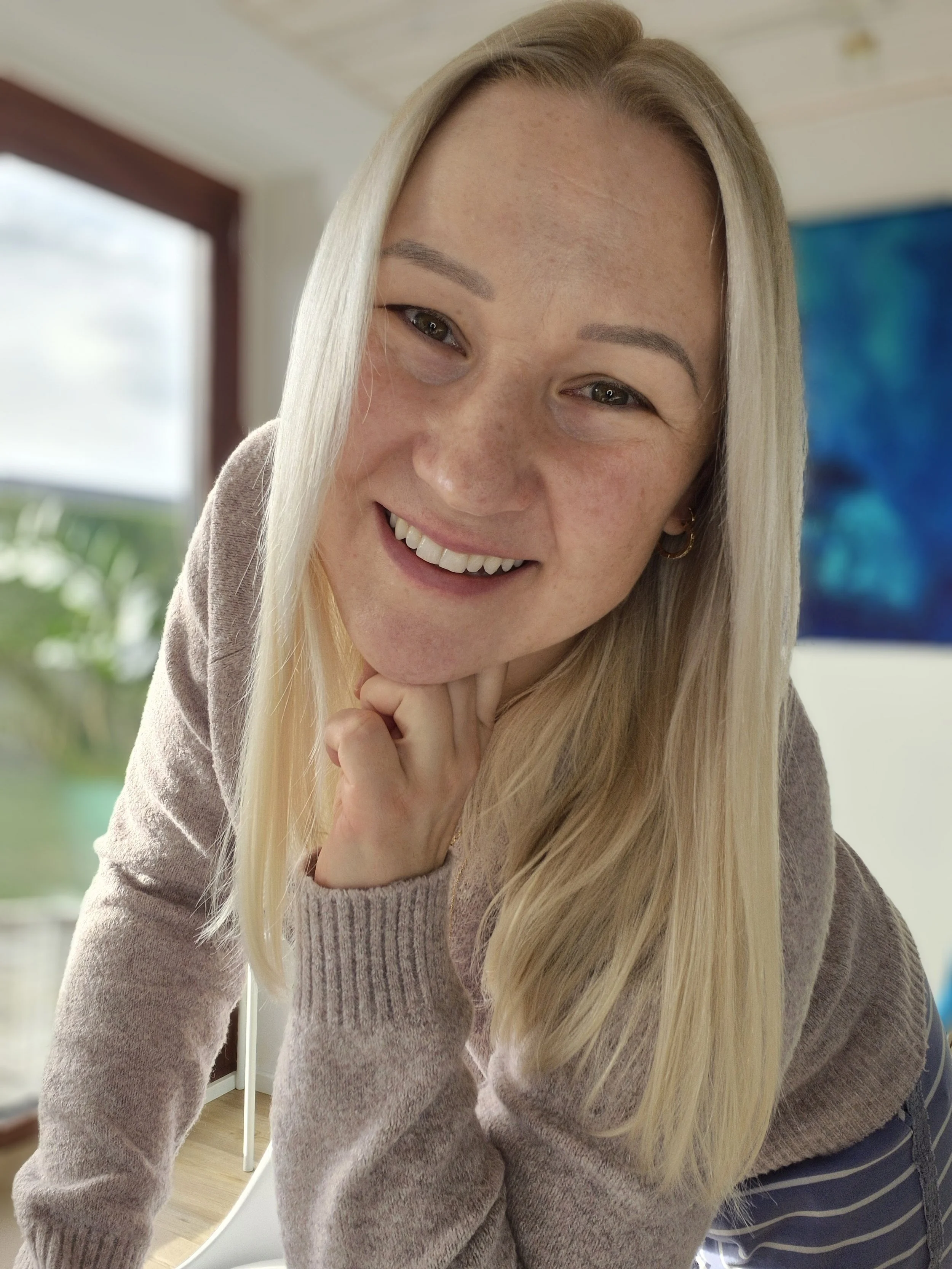 Close-up of a woman smiling, leaning toward the camera with her hand under her chin, inside a brightly lit room with a window and colorful artwork in the background.