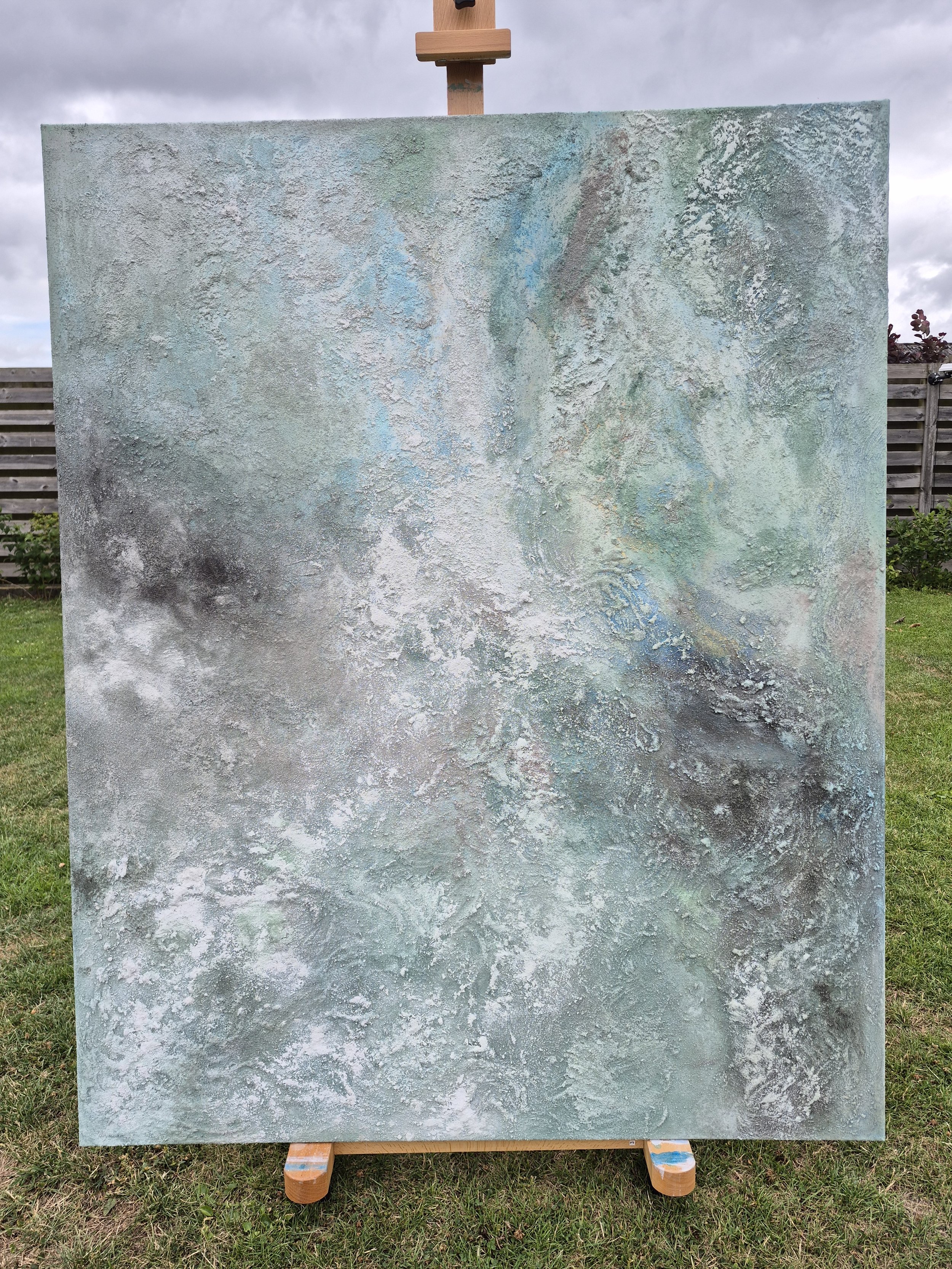 Et meget stort abstrakt maleri med kraftig struktur i hvirvlende lag af teal, søgrøn, grå, kulpigment og hvid. Maleriet hænger på en lys væg i en modern spiseplads over en skænk i lyst træ.