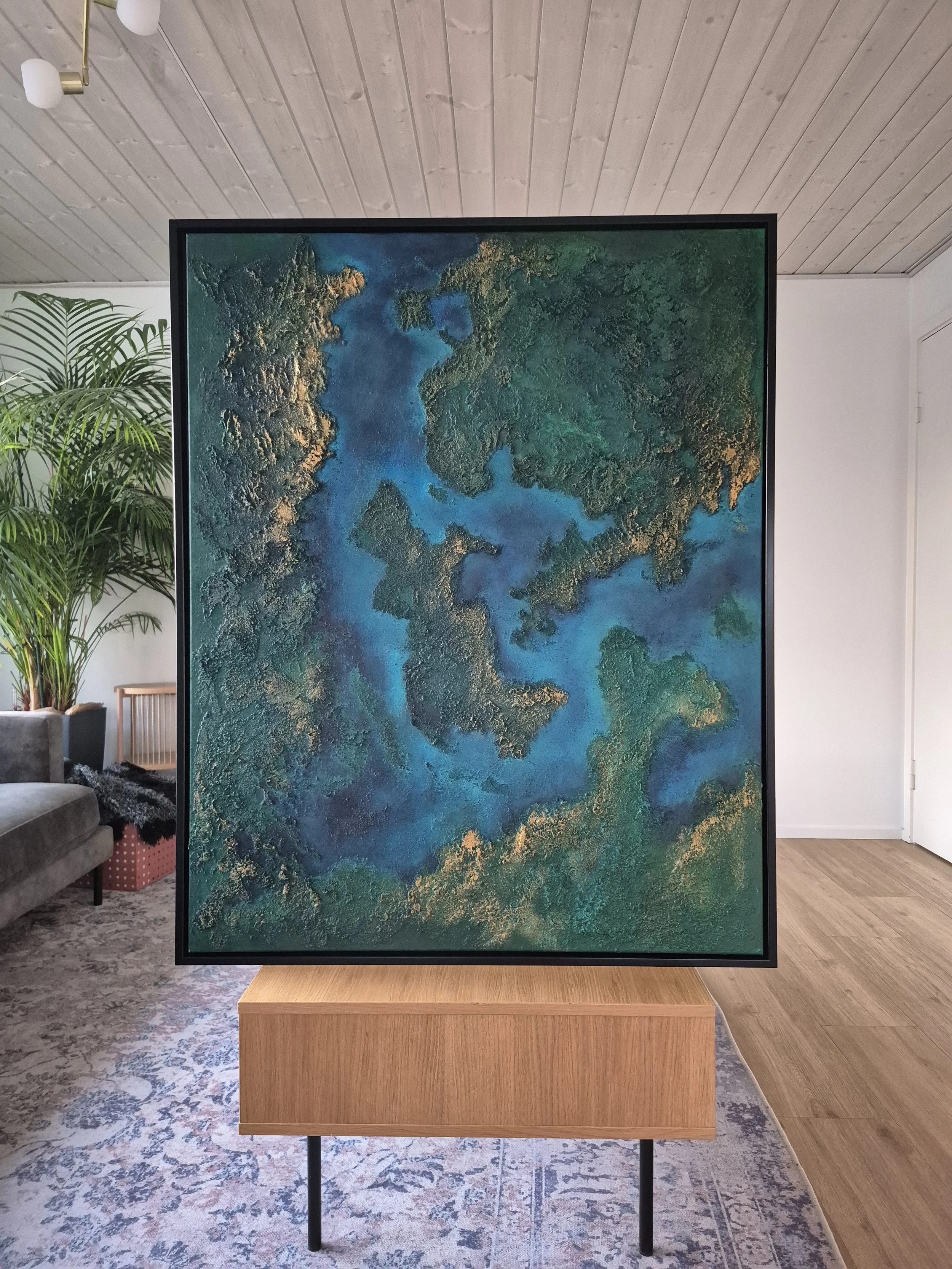 Stort 80x100 cm abstrakt maleri med kraftig tekstur af ægte strandsand. Motivet er i dybe blå, grønne og gyldne nuancer, der ligner et kystlandskab set fra oven.