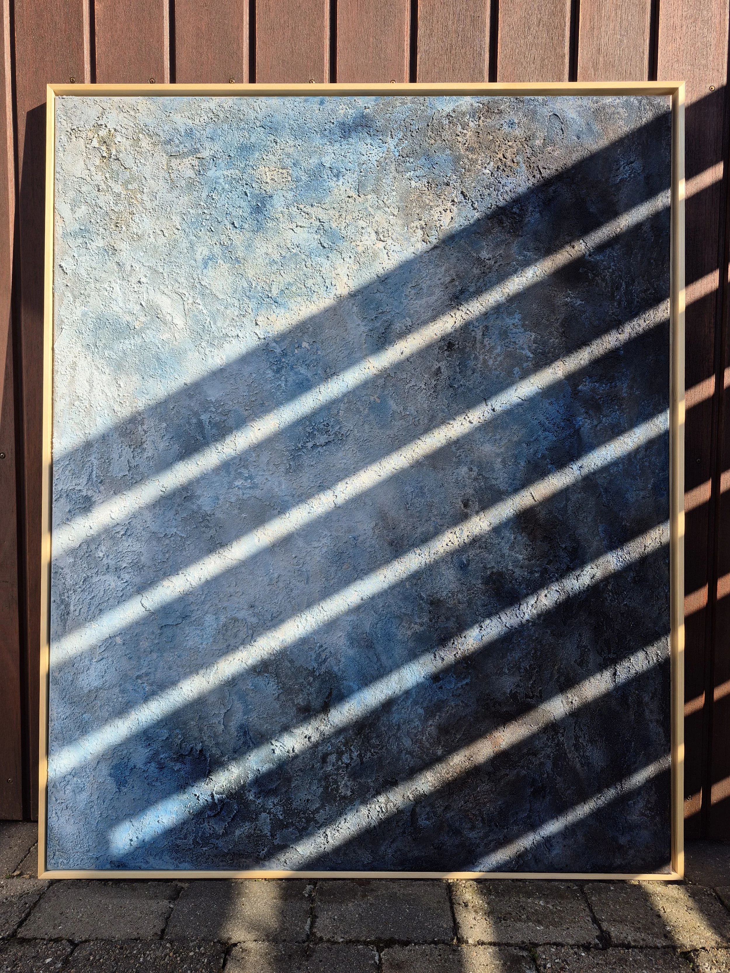 Stort abstrakt maleri med titlen 'Cosmos' udstillet på et staffeli. Maleriet har en kraftig struktur af sand og akryl i blå, grå og beige nuancer, indrammet i en lys træramme.