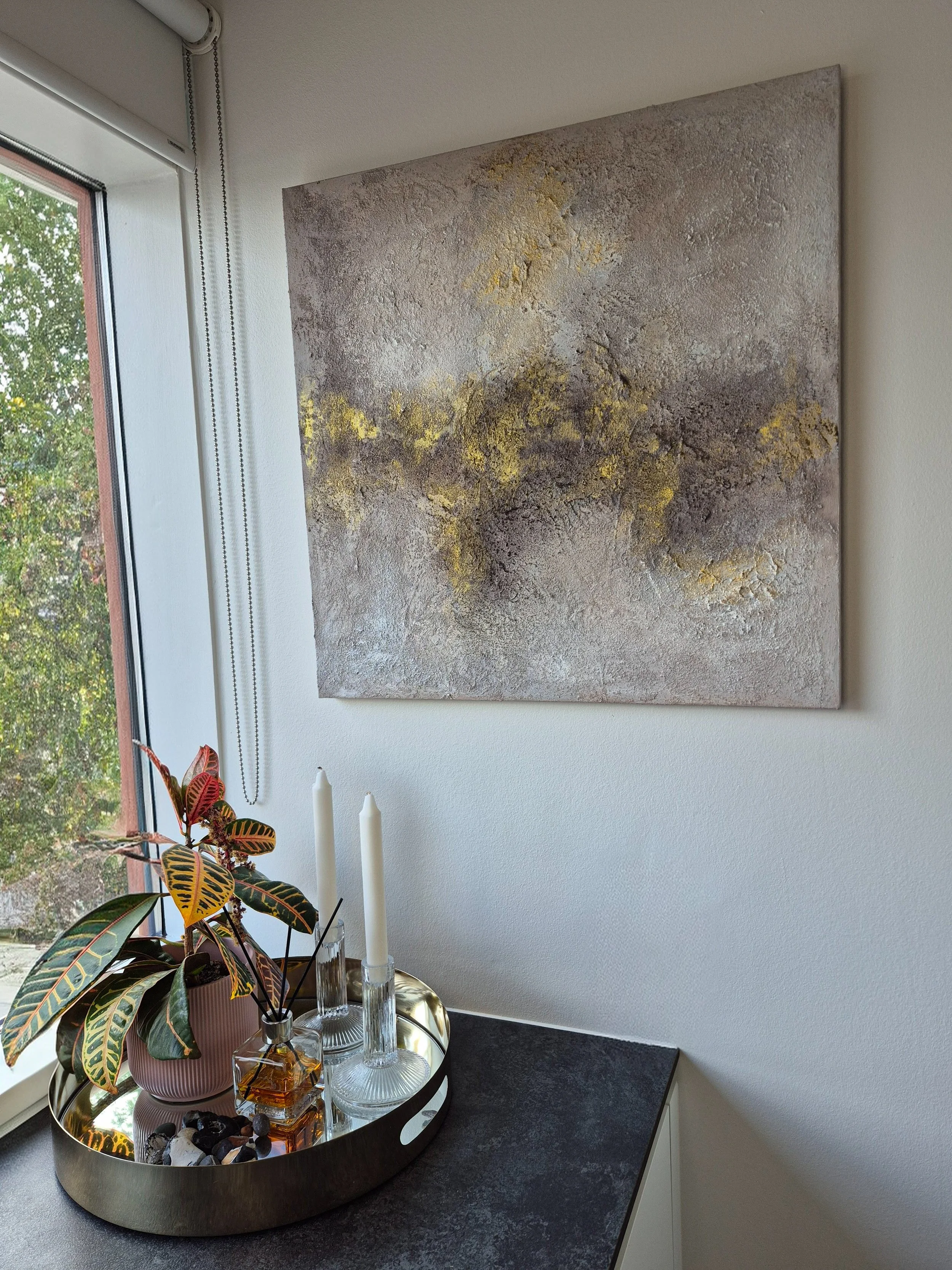 ​A square abstract textured painting featuring earth tones of grey and brown with a vibrant horizontal streak of gold leaf, hung in a bright, modern living room