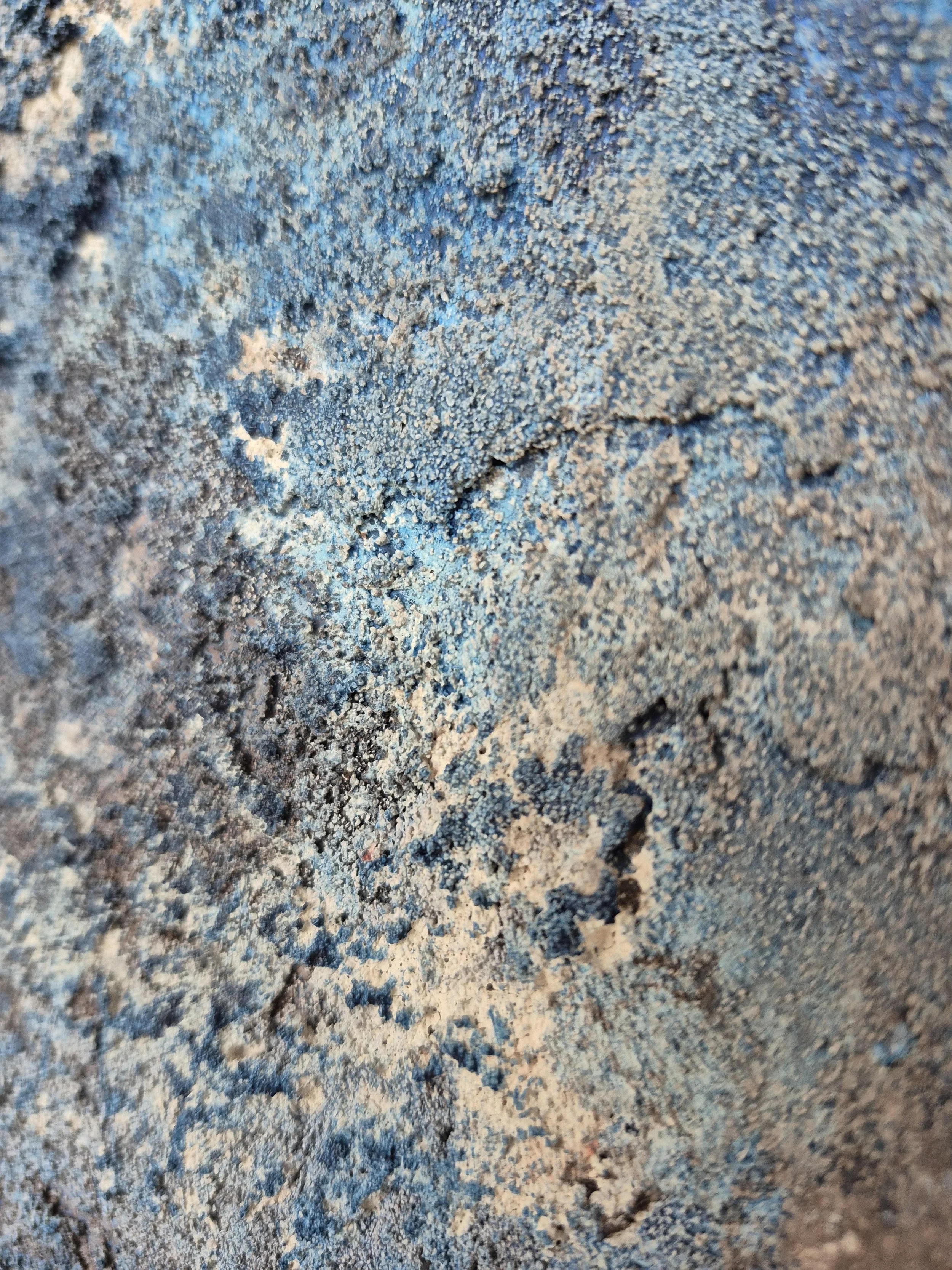 Rundt abstrakt maleri med kraftig tekstur i blå, grå og sorte nuancer, der minder om en planet eller en måneoverflade på en hvid baggrund.