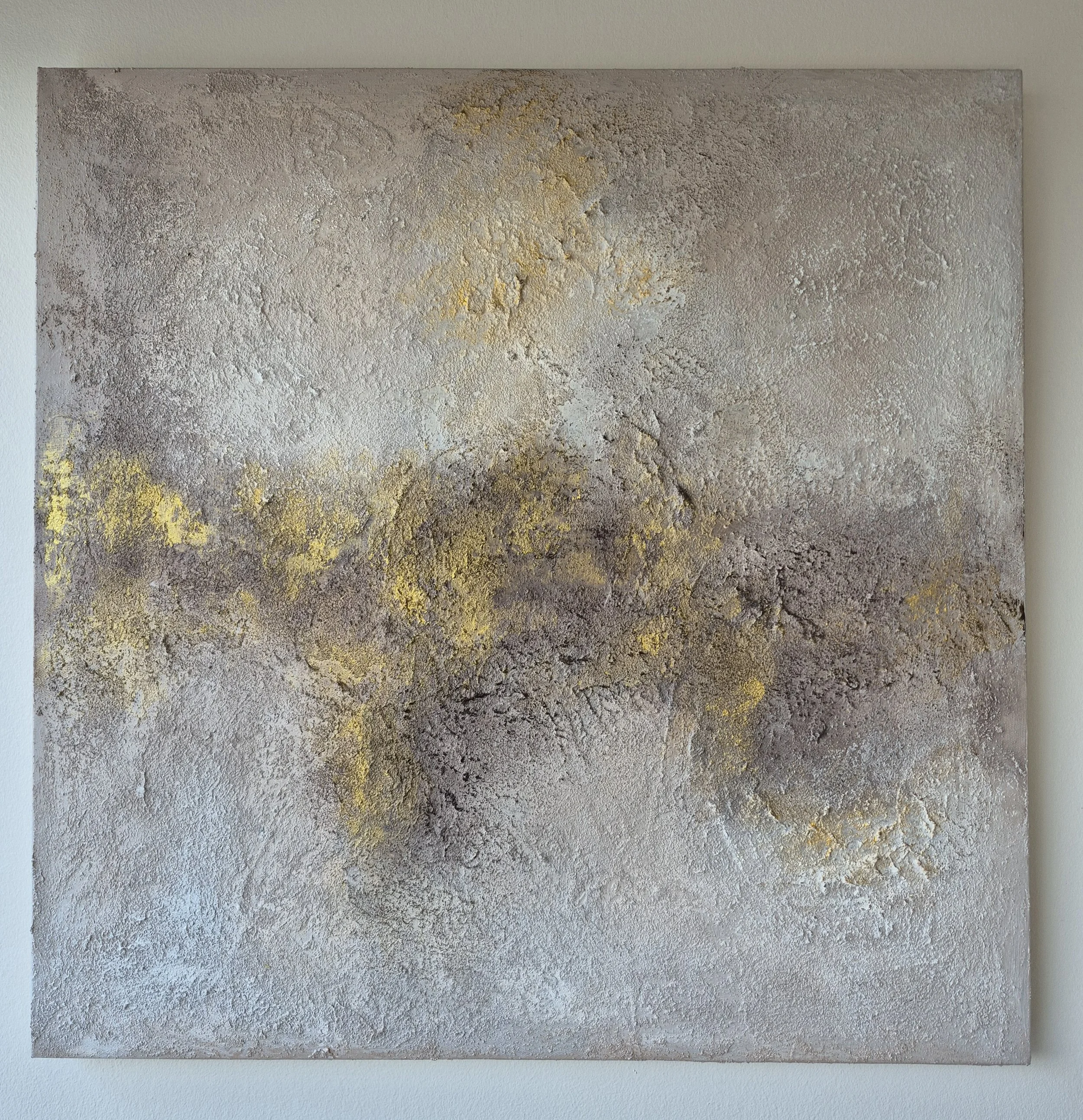 ​A square abstract textured painting featuring earth tones of grey and brown with a vibrant horizontal streak of gold leaf, hung in a bright, modern living room
