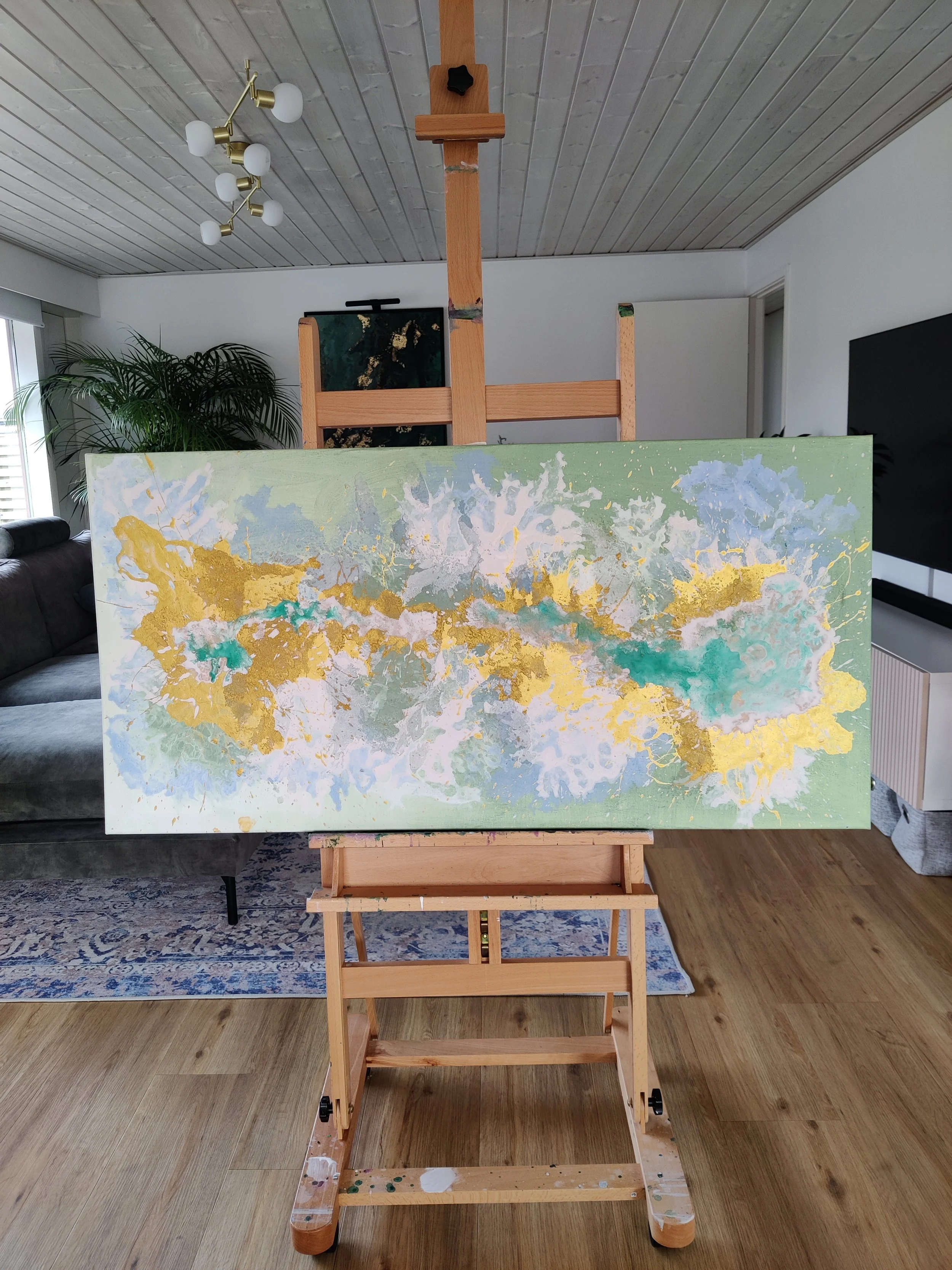 Moderne horisontalt abstrakt maleri i grønne, gyldne og blå nuancer (60x120 cm) over et spisebord i en lys skandinavisk indretning.