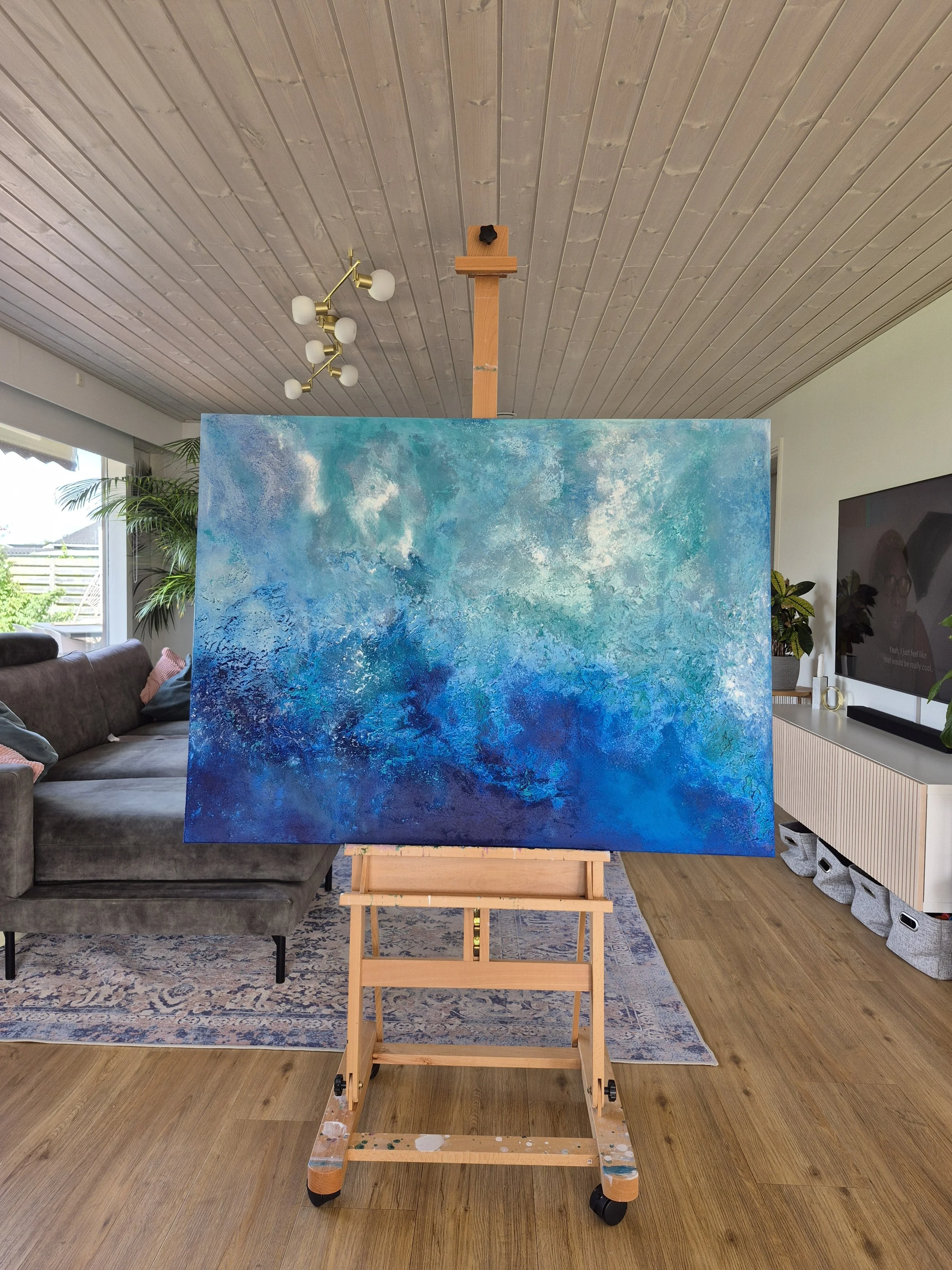 ​Stort blåt abstrakt maleri (90x120 cm) med nuancer af turkis, navy og hvid monteret i en moderne lys stue. Moderne dansk kunst til væggen.