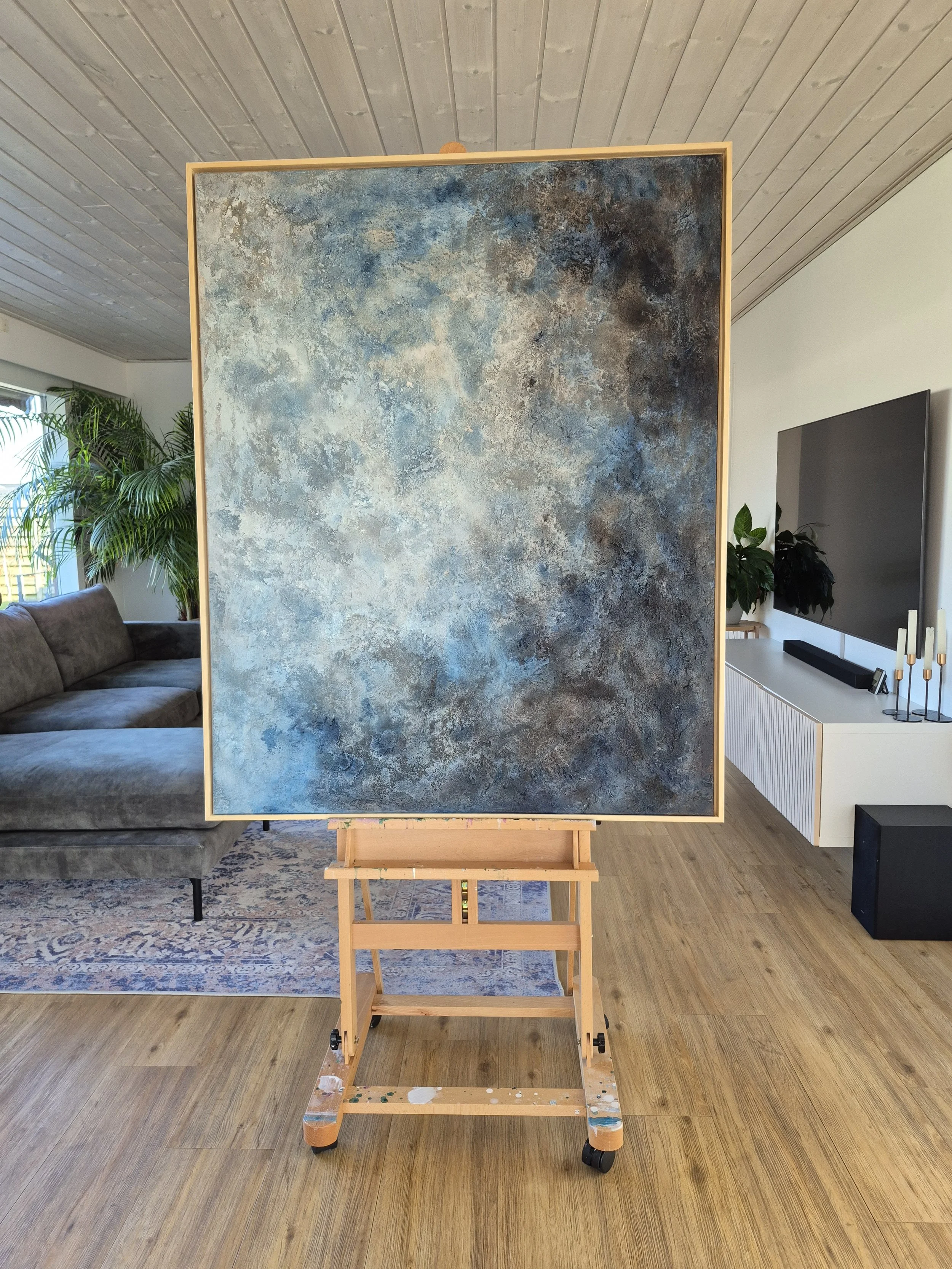 Stort abstrakt maleri med titlen 'Cosmos' udstillet på et staffeli. Maleriet har en kraftig struktur af sand og akryl i blå, grå og beige nuancer, indrammet i en lys træramme.