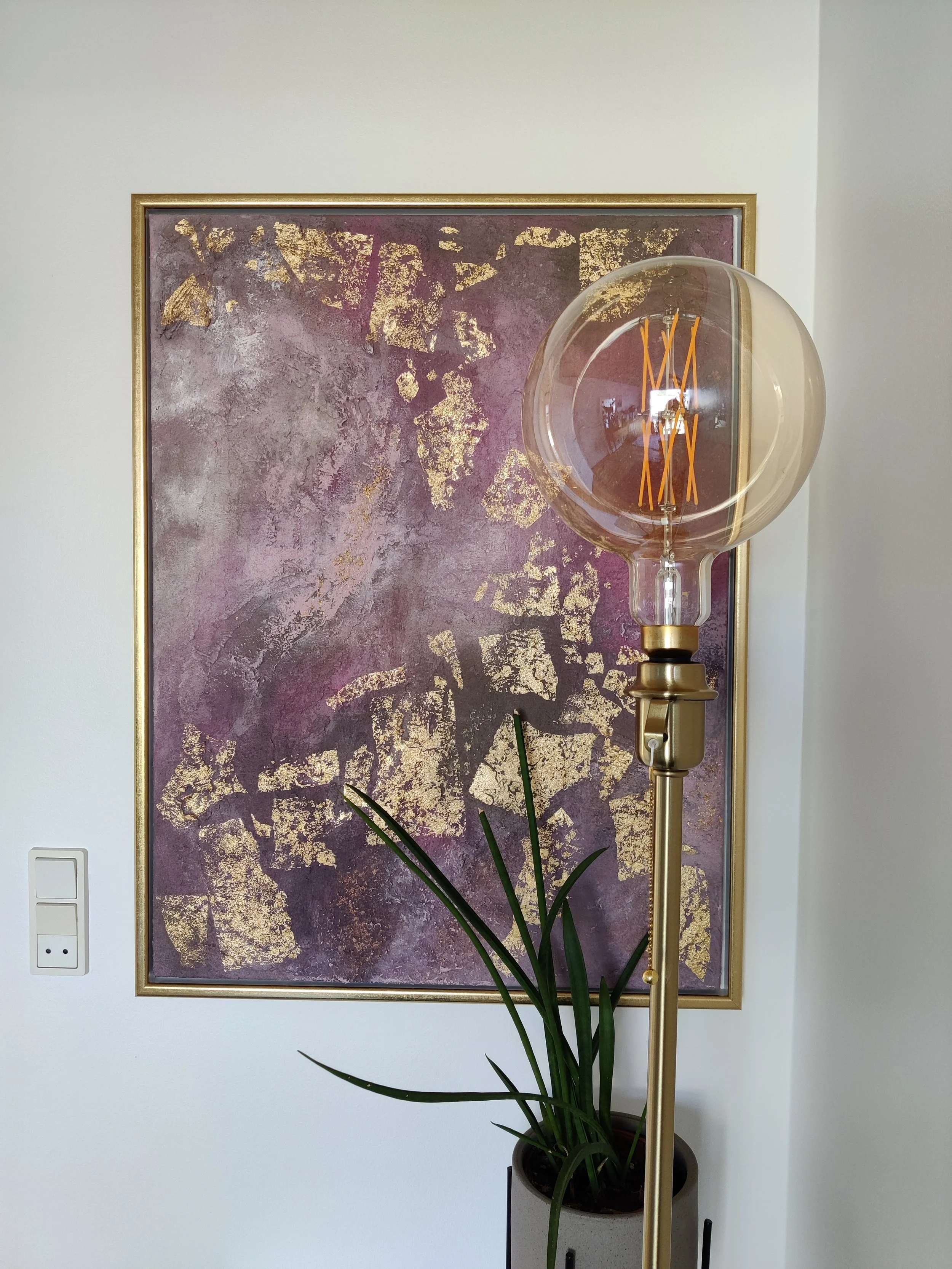 Abstrakt maleri i lilla og rosa nuancer med grov sandstruktur og store guldflager i en smal guldramme.