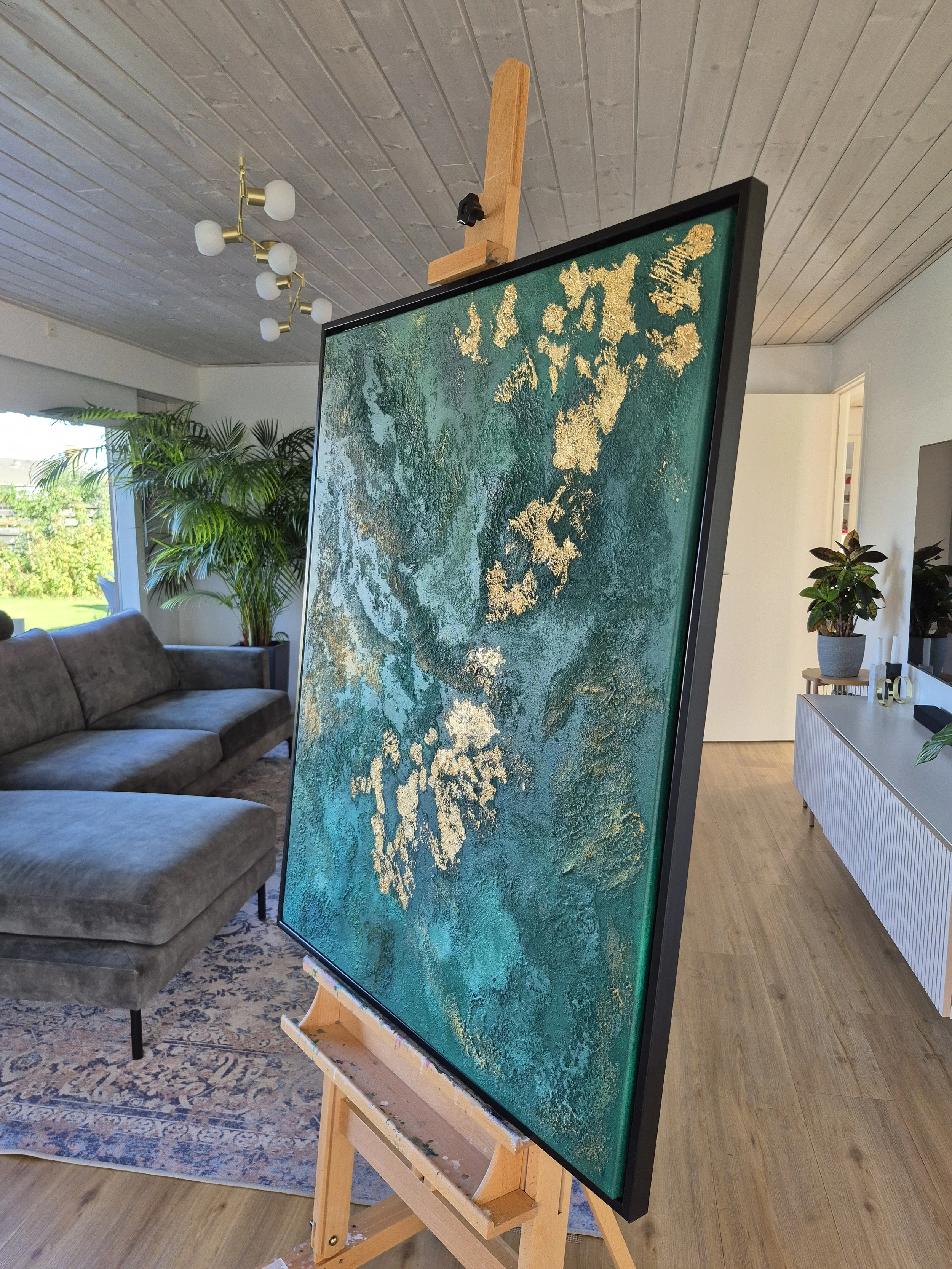 Abstrakt maleri i grønne og gyldne farver 80x100 cm over en grøn sofa i en stue med skandinavisk design.