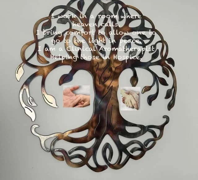 Decorative wall art of a tree made of metal with two photos of hands, one darker skin and one lighter skin, integrated into the design. Overlaid text reads: "I work in a room where heaven calls. I bring comfort to allow one to go to the light in peace. I am a Clinical Aromatherapist Helping those in Hospice."