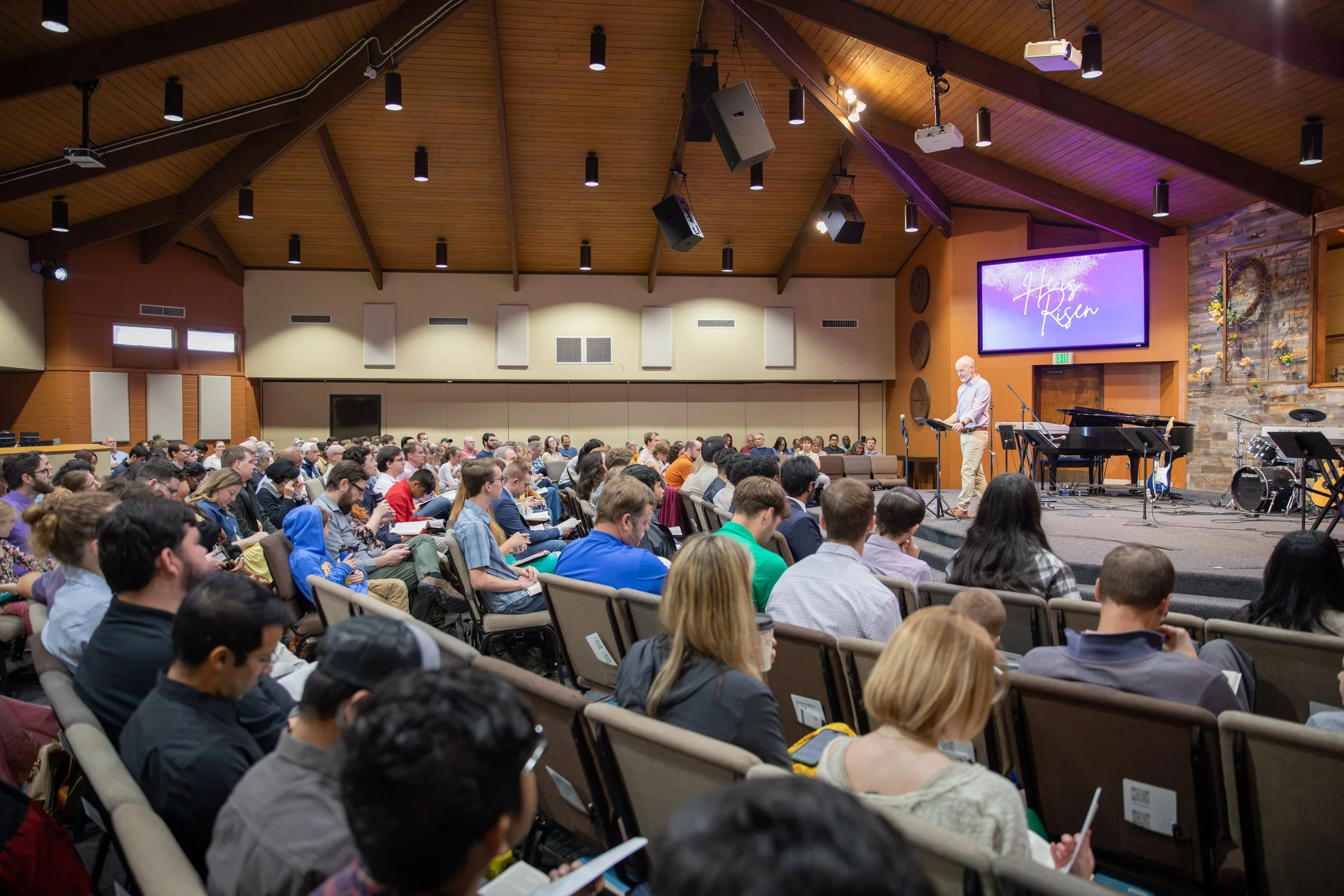A large audience seated in rows watching a speaker on stage in a church-like setting. The stage has musical instruments and a large screen displaying the words 'Haby Rsem' with a purple background.