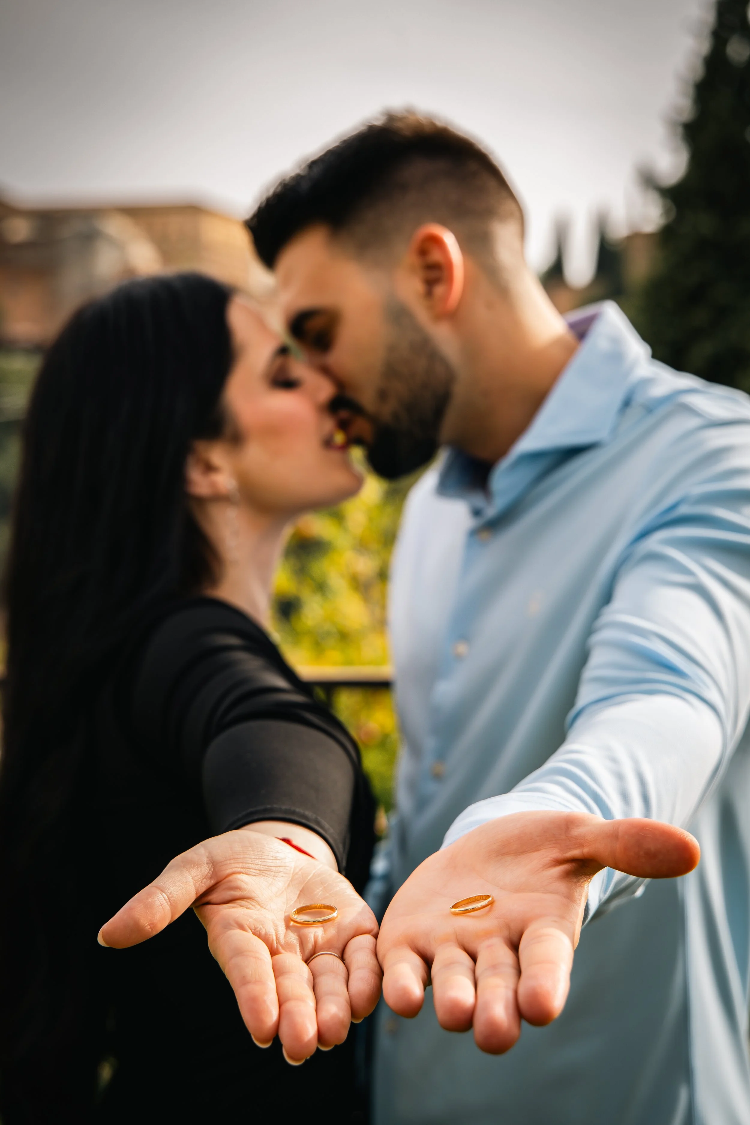 A couple kissing outdoors holding wedding rings in their palms toward the camera.