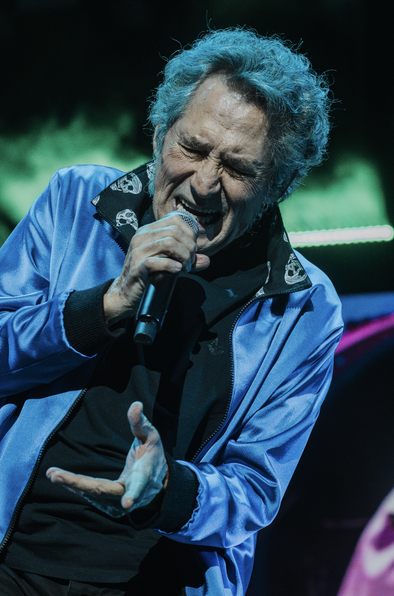 An elderly man with gray, curly hair singing passionately into a microphone on stage, wearing a blue jacket with a skull pattern on the collar.
