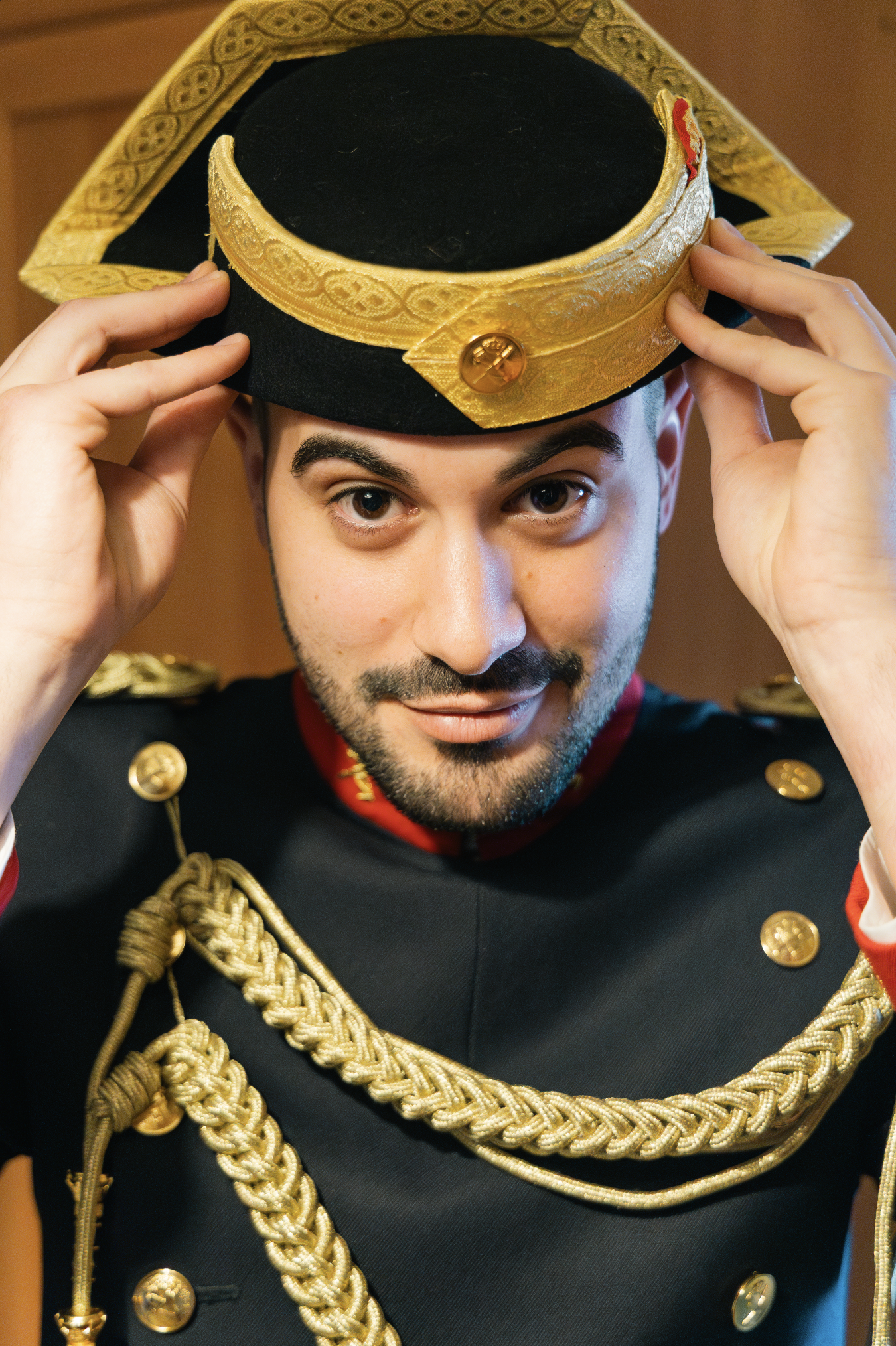 A man dressed in a military-style uniform with gold braid, buttons, and decorations, adjusting a decorative hat with gold trim and emblem.