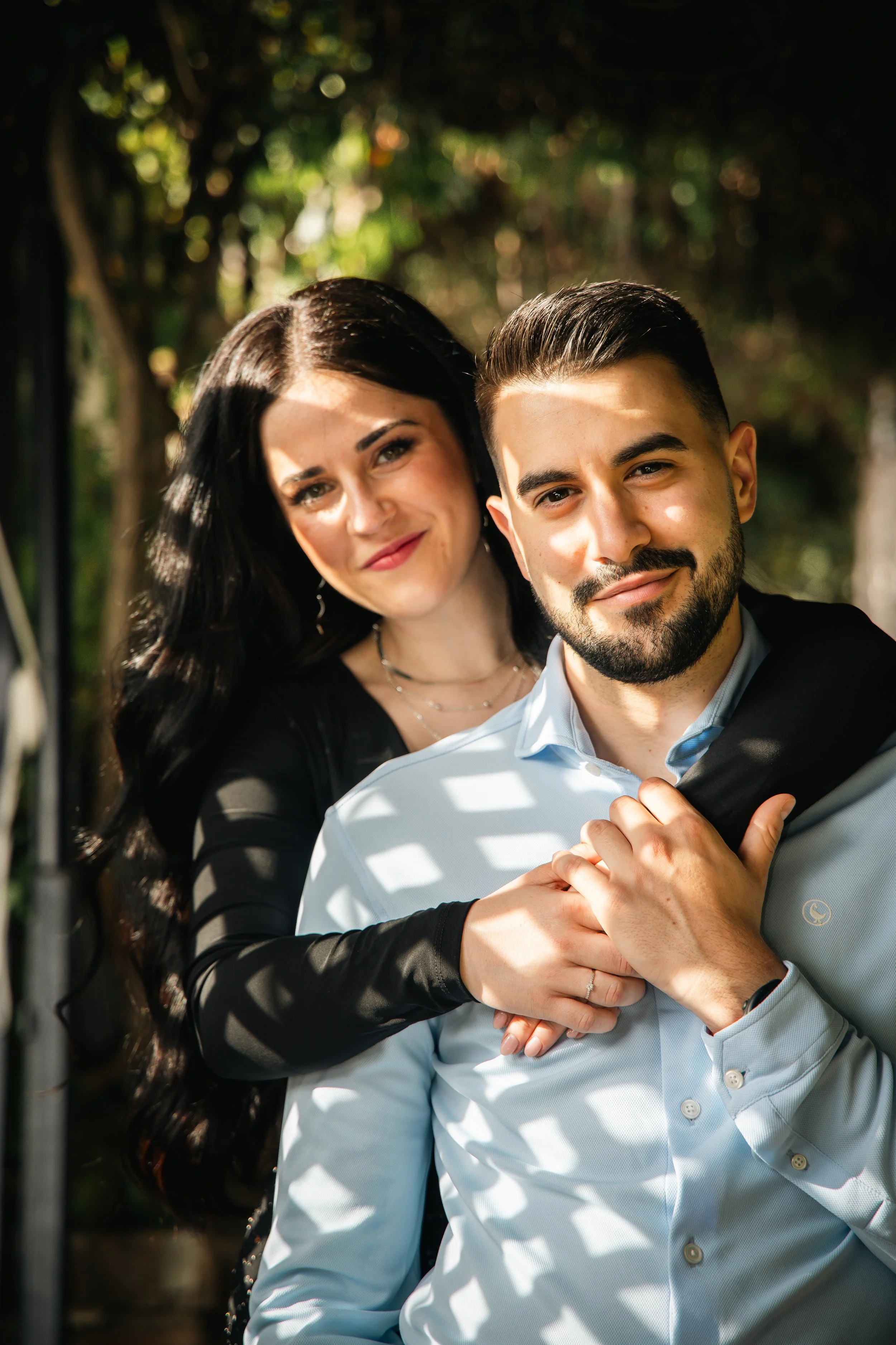 A woman with long black hair and a man with dark hair and beard posing outdoors with sunlight filtering through trees.
