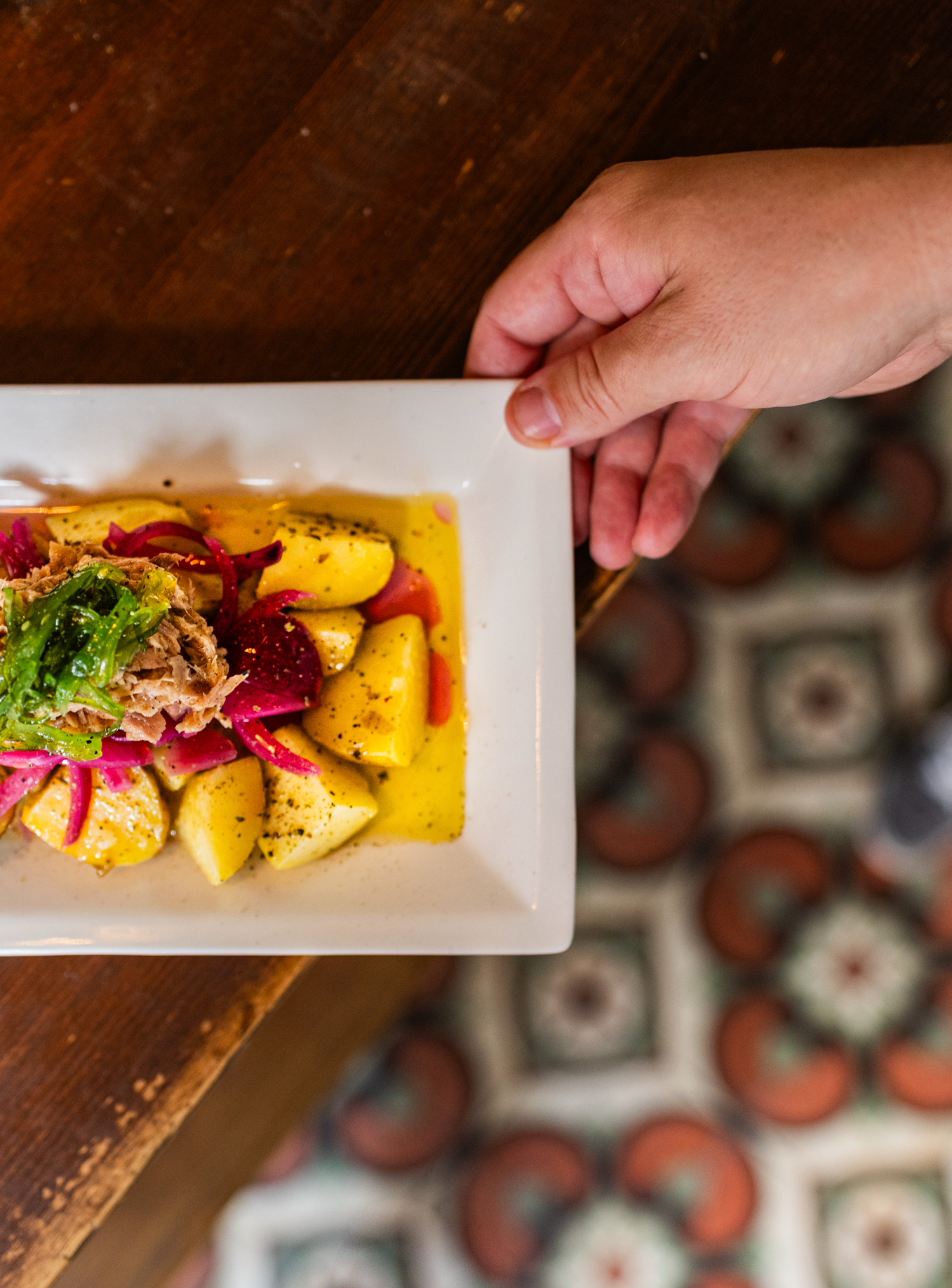 A person holding a white square plate with sliced potatoes, pickled red onions, shredded meat, and herbs in a yellow sauce. The setting includes a wooden table and colorful patterned floor tiles.