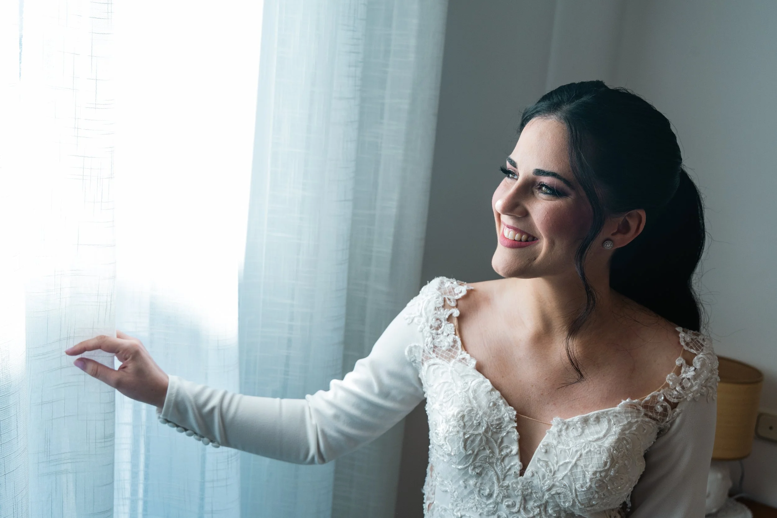 A smiling bride with dark hair in a low ponytail, wearing a white wedding dress with lace and embroidery details, looking out of a window with sheer curtains.