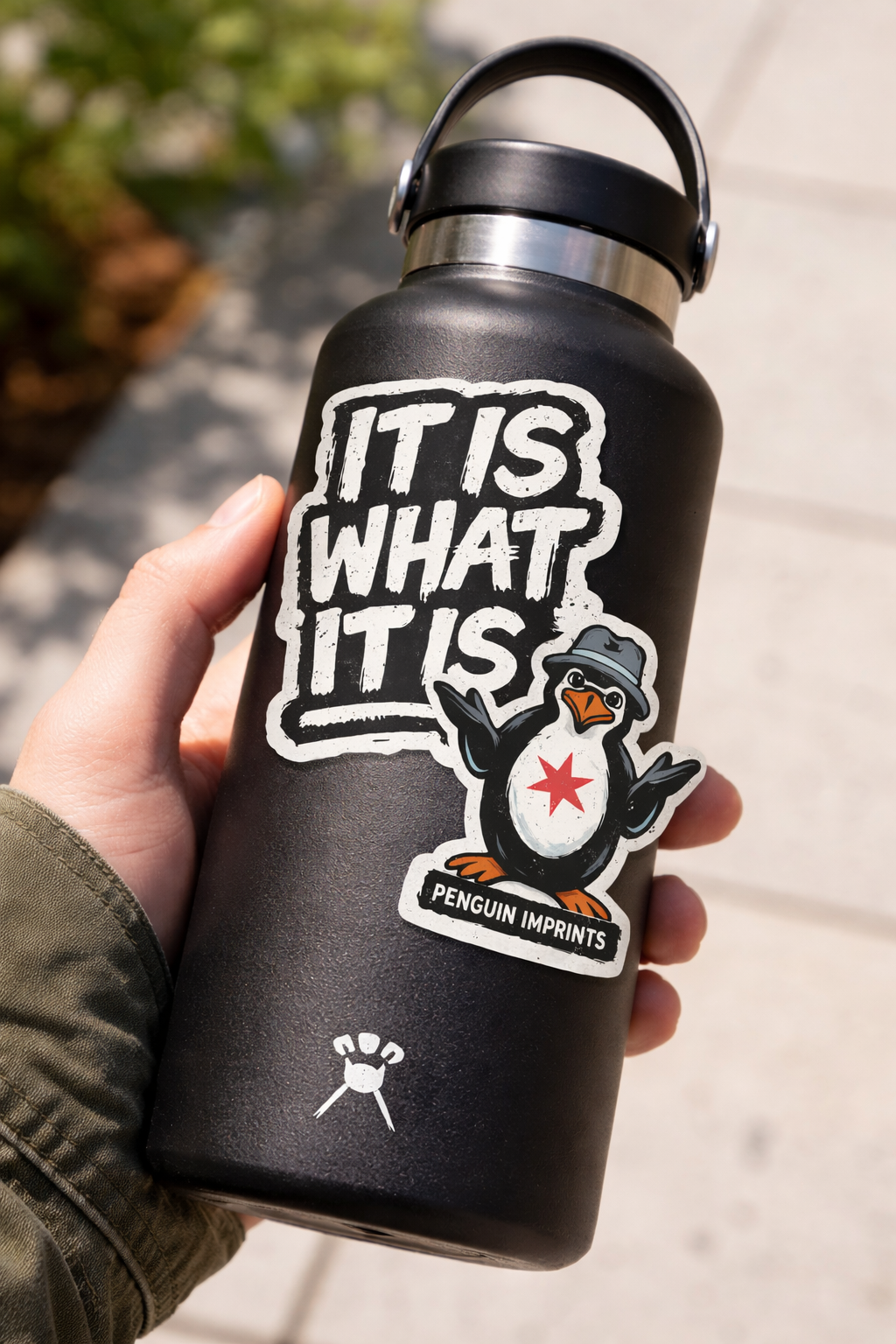 A black water bottle with a sticker featuring the phrase 'It is what it is' and a cartoon penguin wearing a gray fedora and a red star on its chest, with the words 'Penguin Imprints' underneath.
