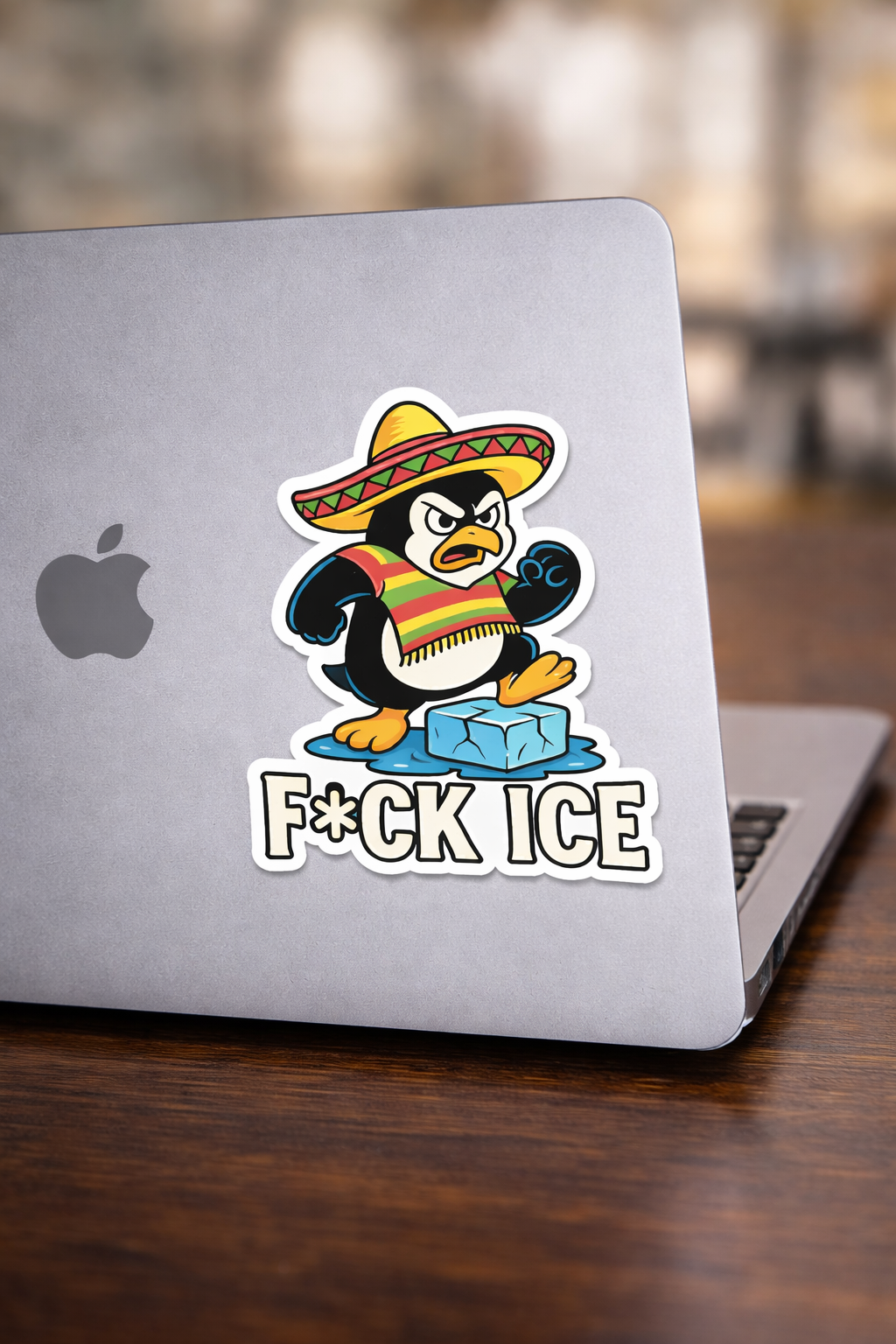 A sticker on a silver Apple laptop features a cartoon penguin wearing a Mexican sombrero and a colorful poncho, standing on an ice block with water around it. The text on the sticker says "F*CK ICE."