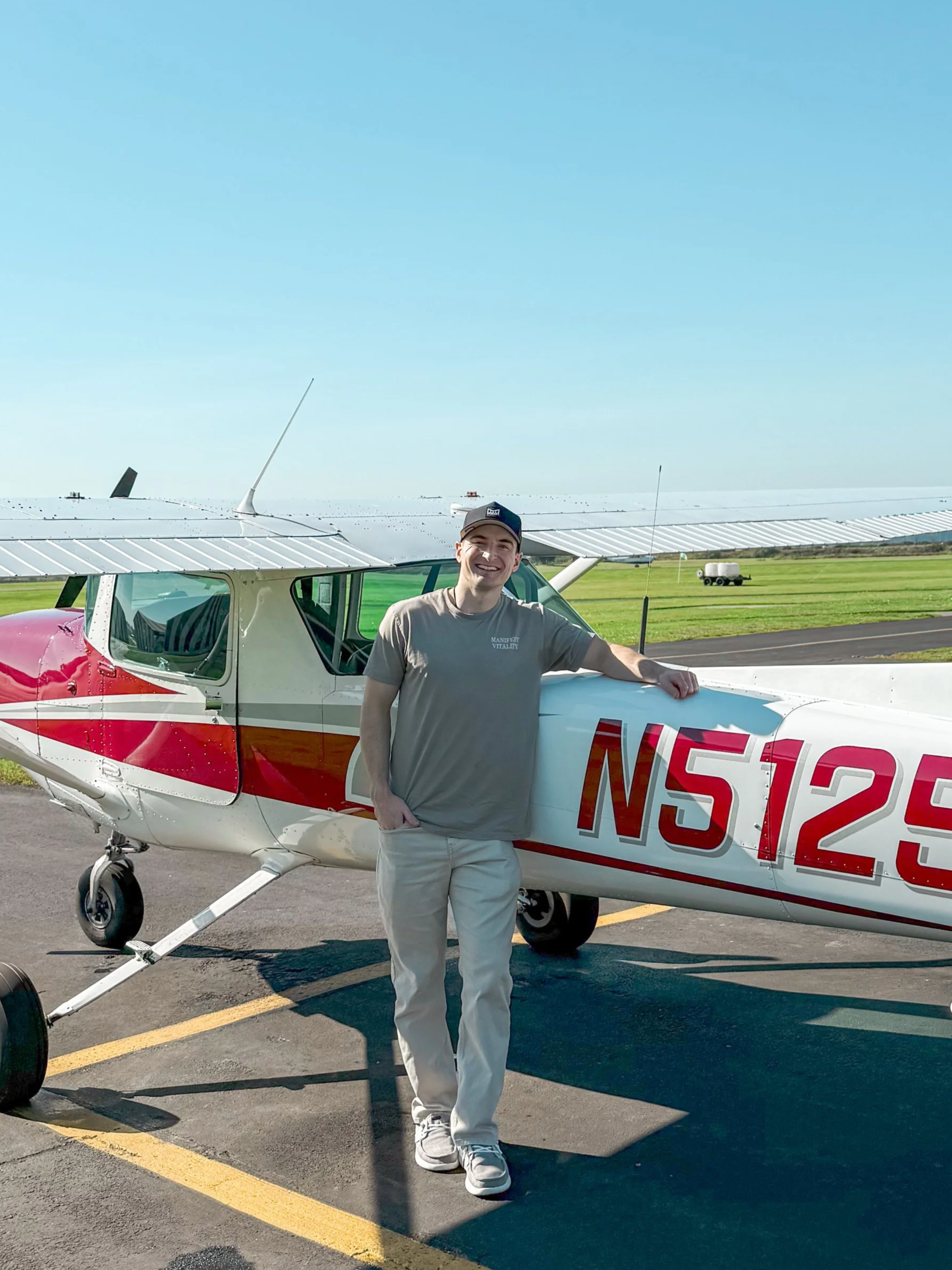 A young man smiling and leaning on a small airplane with the registration N5125, parked on an airport tarmac on a sunny day.