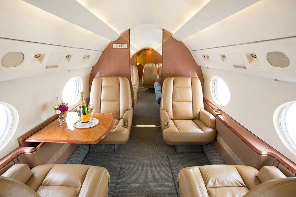 Interior of an airplane cabin with beige leather seats, small round windows, a wooden table with a champagne bottle and flowers, and an emergency exit sign.