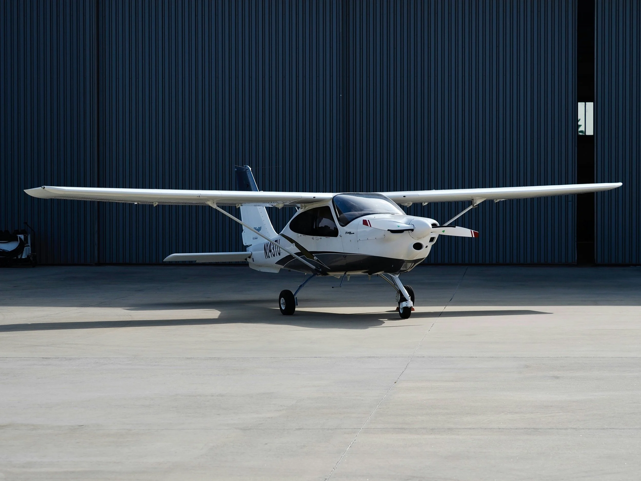 Small white airplane parked on tarmac in front of a large blue hangar.