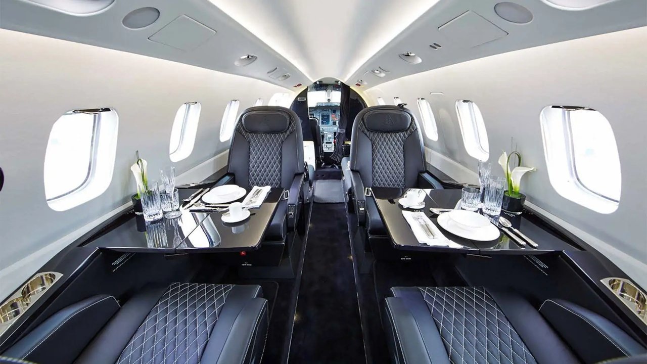 Interior of a private jet with two luxury seats, a dining table set with plates, glasses, and utensils, and windows along the side.