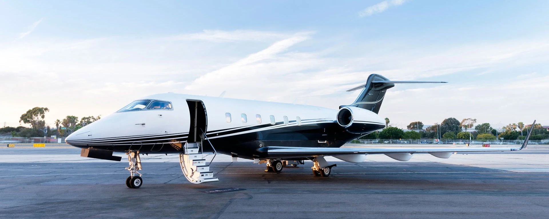 Private jet airplane on airport tarmac with stairs at the open door, trees and a partly cloudy sky in the background.