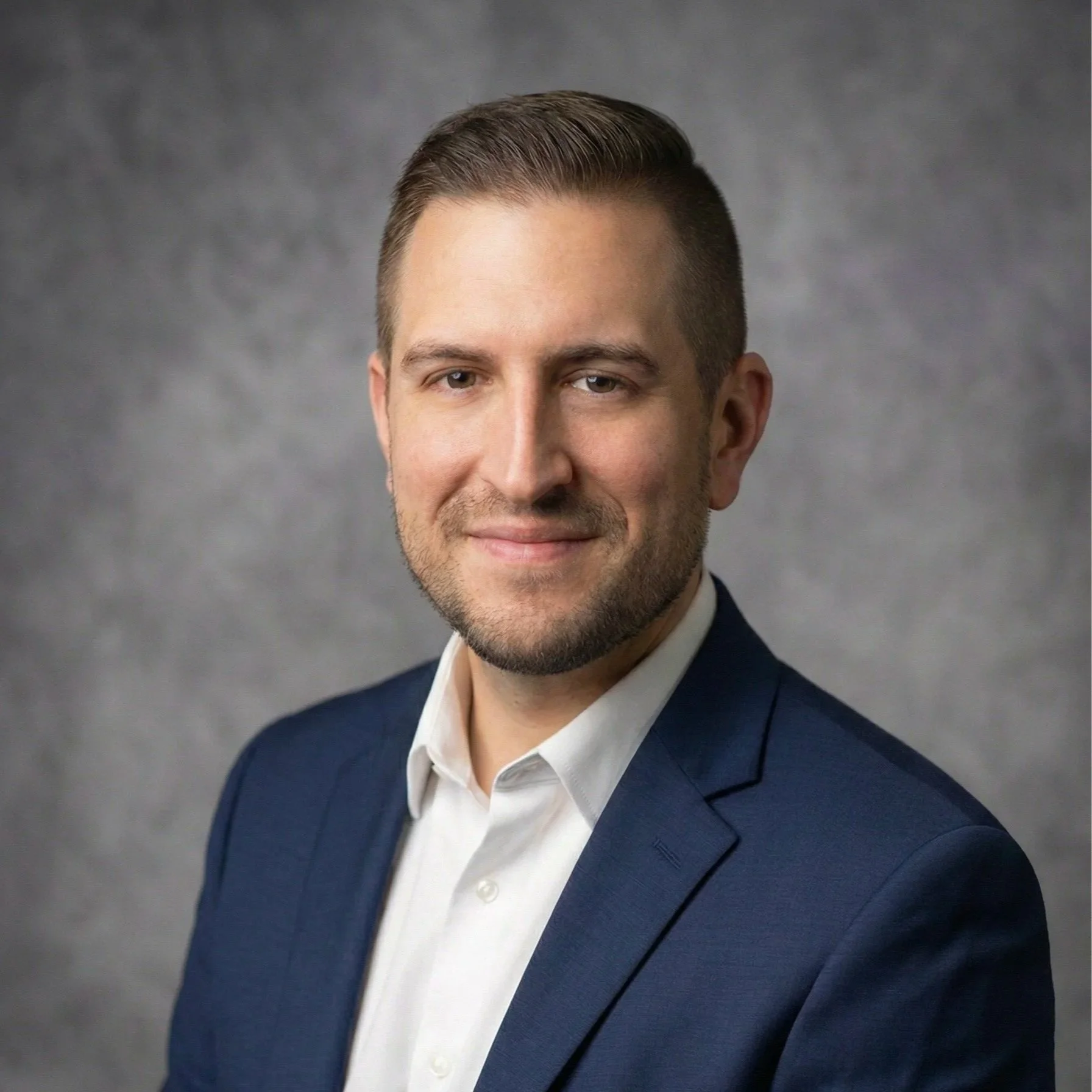 Headshot of a man with light skin, short brown hair, and a beard, wearing a navy blazer and white shirt, smiling against a gray textured background.