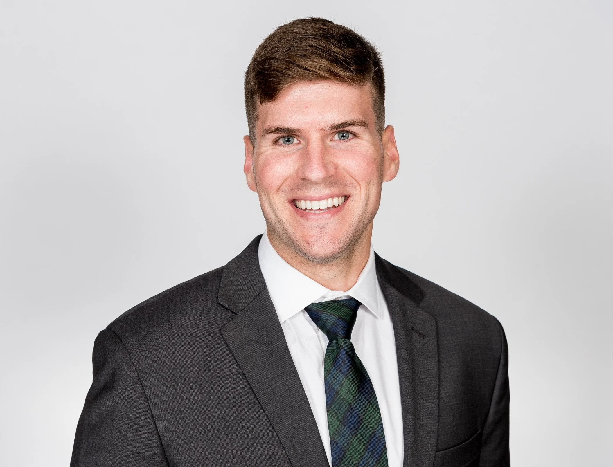 Headshot of a young man in a black suit, white shirt, and a plaid tie, smiling against a neutral background.
