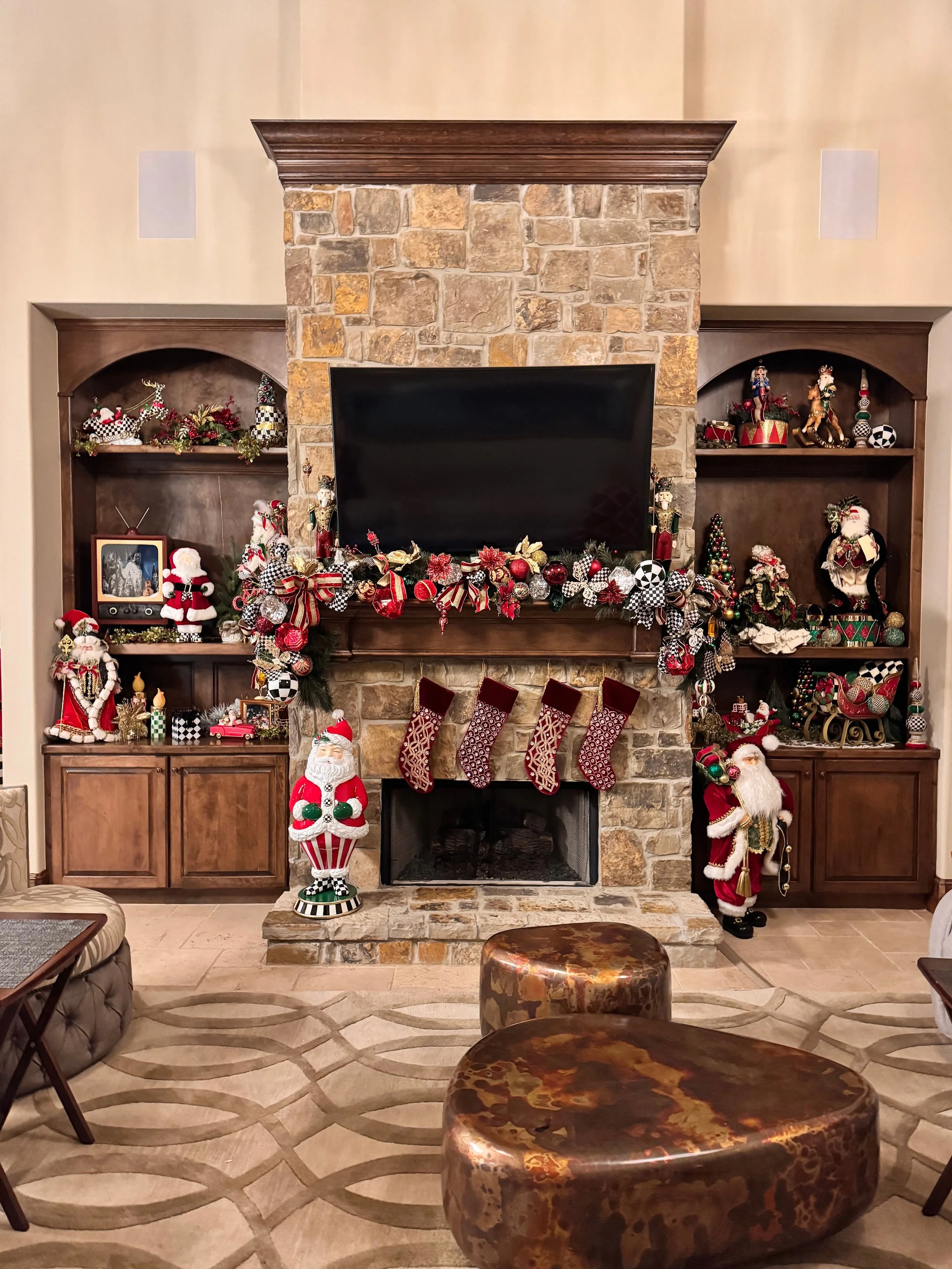 Living room decorated with Christmas decorations on a wooden hearth, including stockings, Santa figurines, and garlands, with built-in shelves on either side filled with holiday ornaments and figures, and a television mounted above the fireplace.