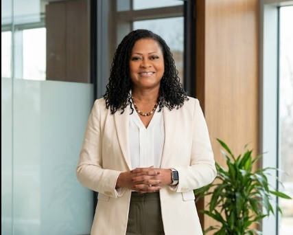 A woman smiling, wearing a cream blazer over a white blouse, standing in an office with large windows and a plant in the background.