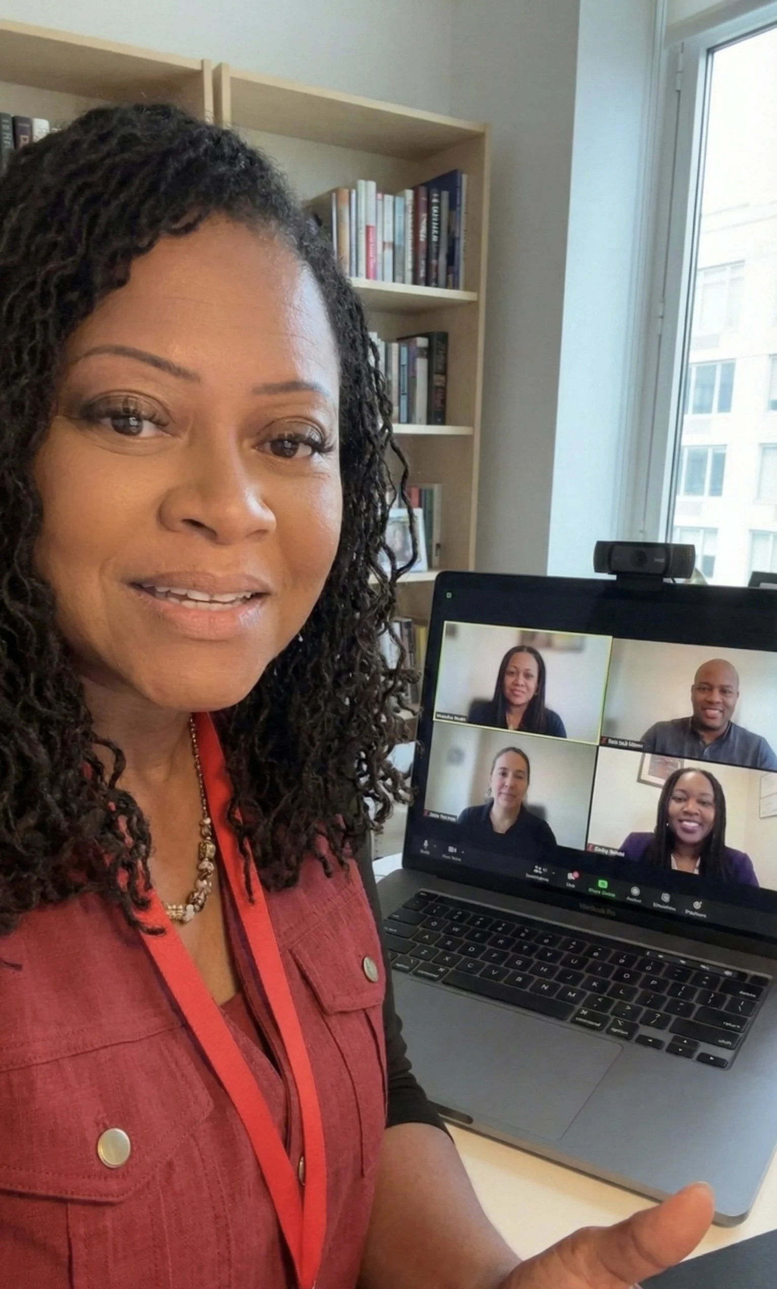 A woman with dark curly hair taking a selfie during a virtual meeting on her laptop. The screen shows four participants in the video call, two women and two men, with a bookshelf and window in the background.