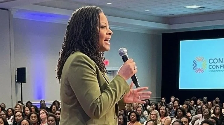 Woman speaking into a microphone at a conference with a large audience and a conference logo on a screen in the background.