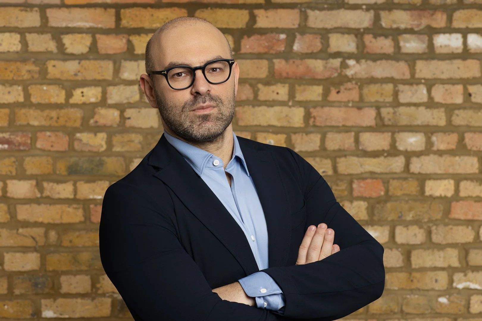 A man with glasses, a beard, and a bald head is standing with arms crossed in front of a brick wall. He is wearing a dark blazer over a light blue shirt.