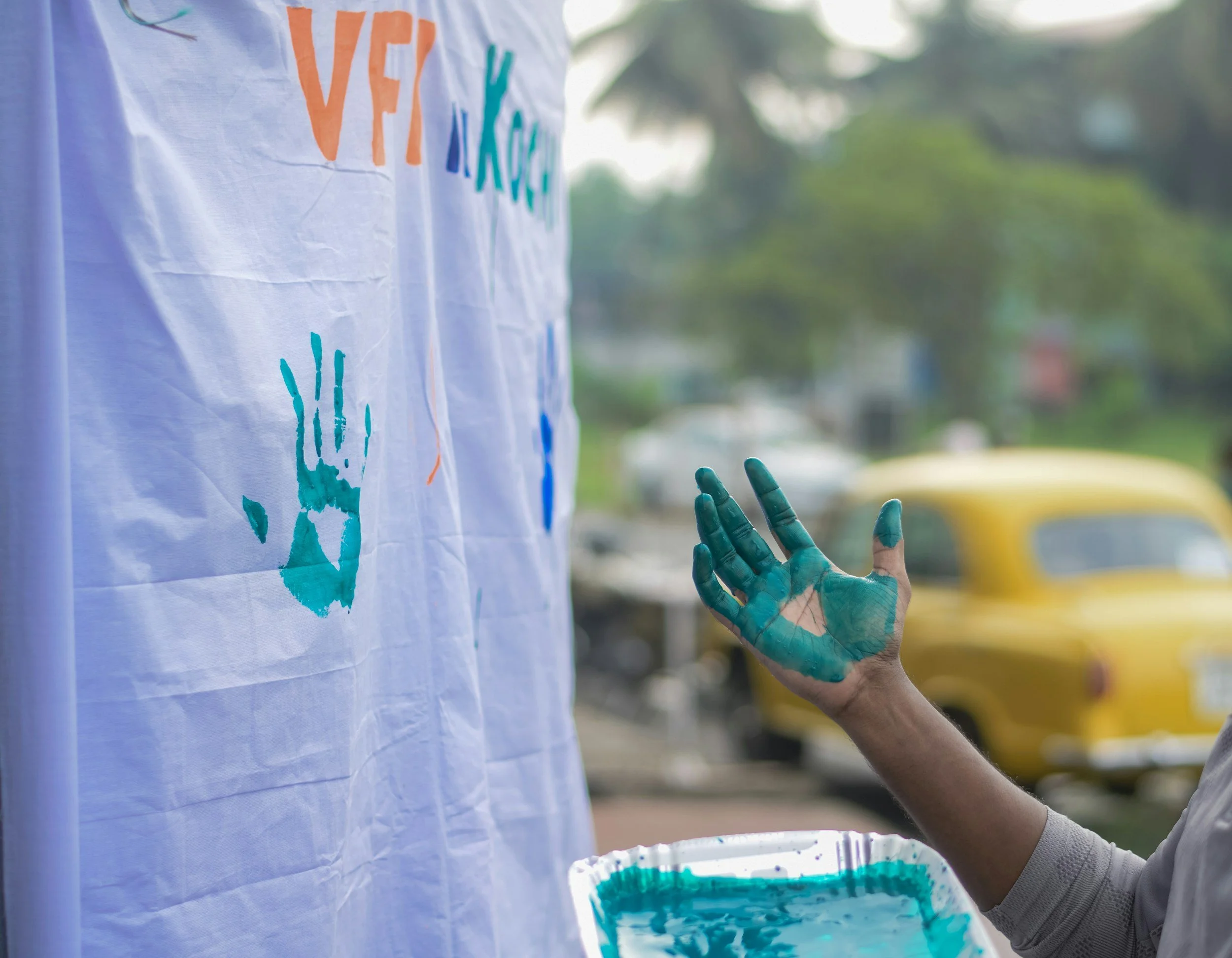 A person with a painted hand reaching out next to a cloth with colorful handprints and text, holding a tray of green paint.