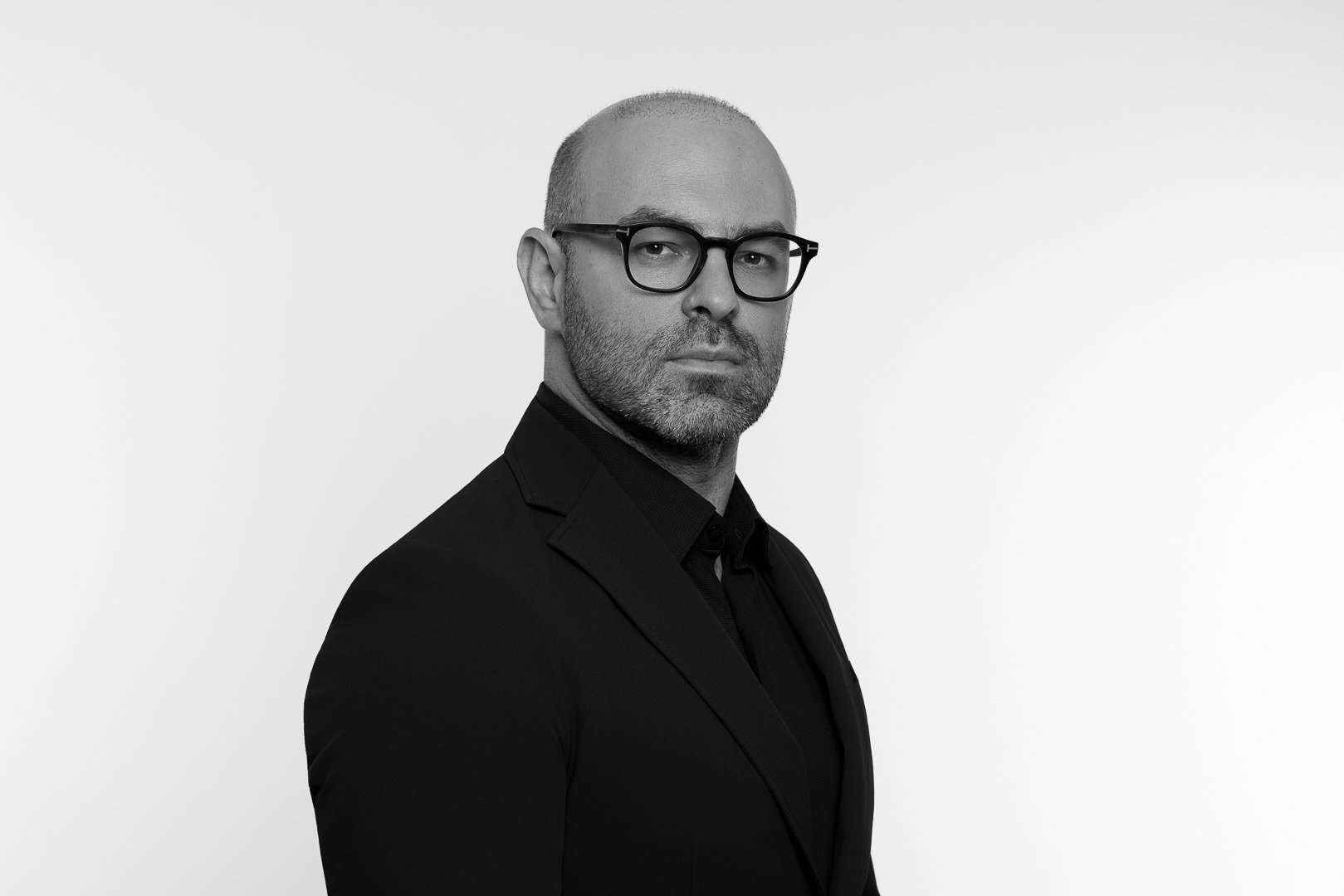 Black and white portrait of a man with glasses, short hair, and a beard, wearing a dark suit against a plain white background.