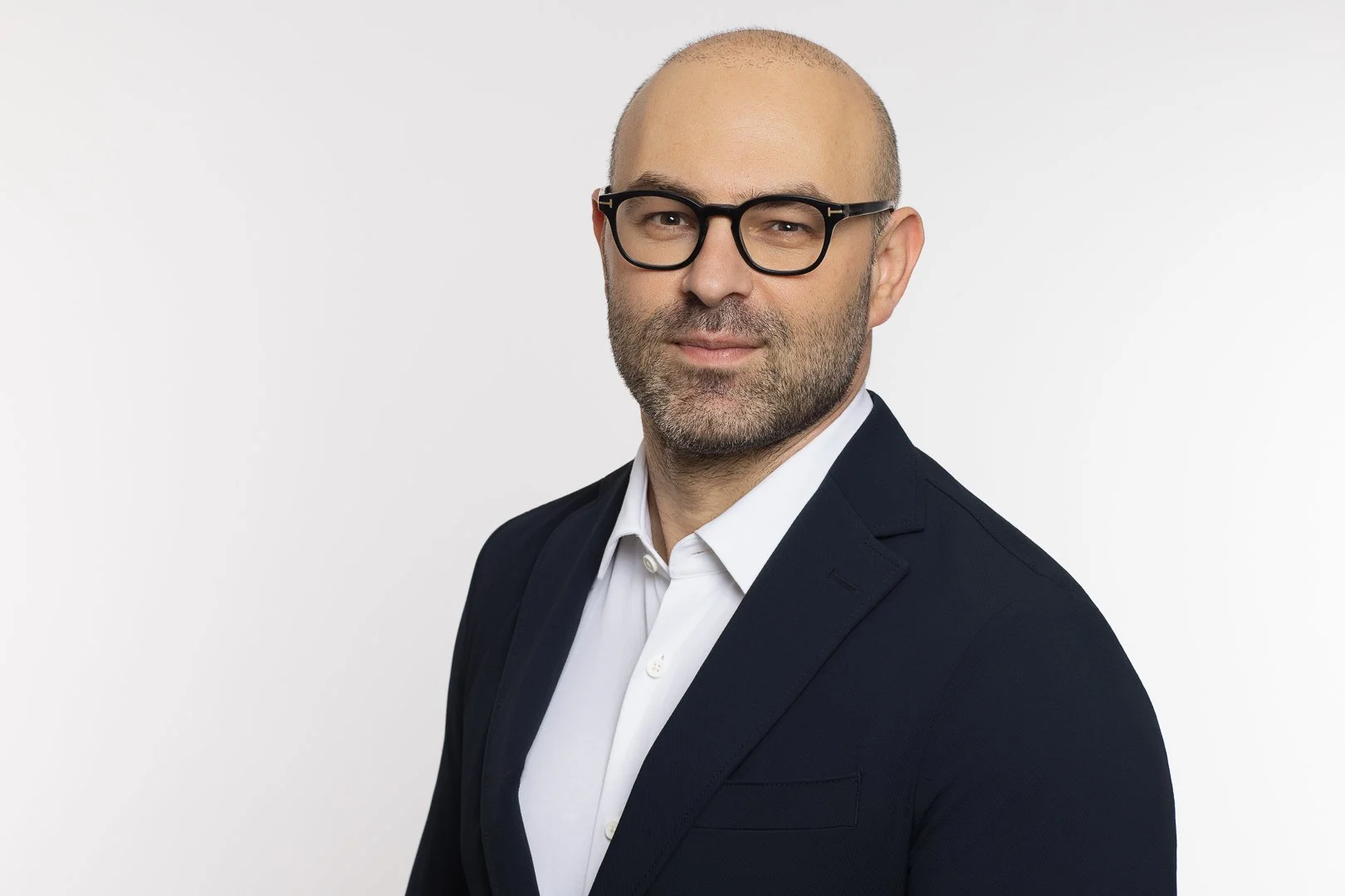 A man with a bald head, glasses, and a beard wearing a dark suit and white shirt, posing against a plain white background.