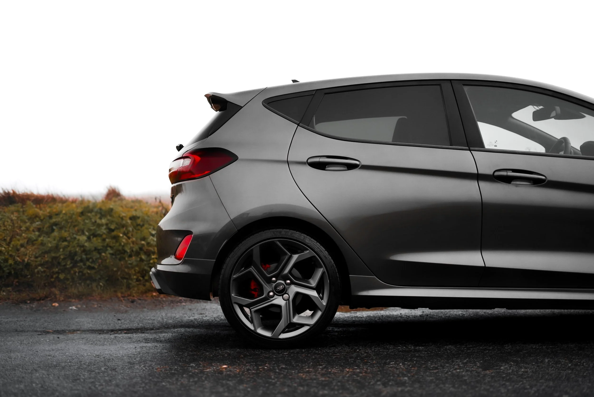 Close-up of a gray modern hatchback car parked on a wet asphalt surface with a background of bushes and overcast sky.