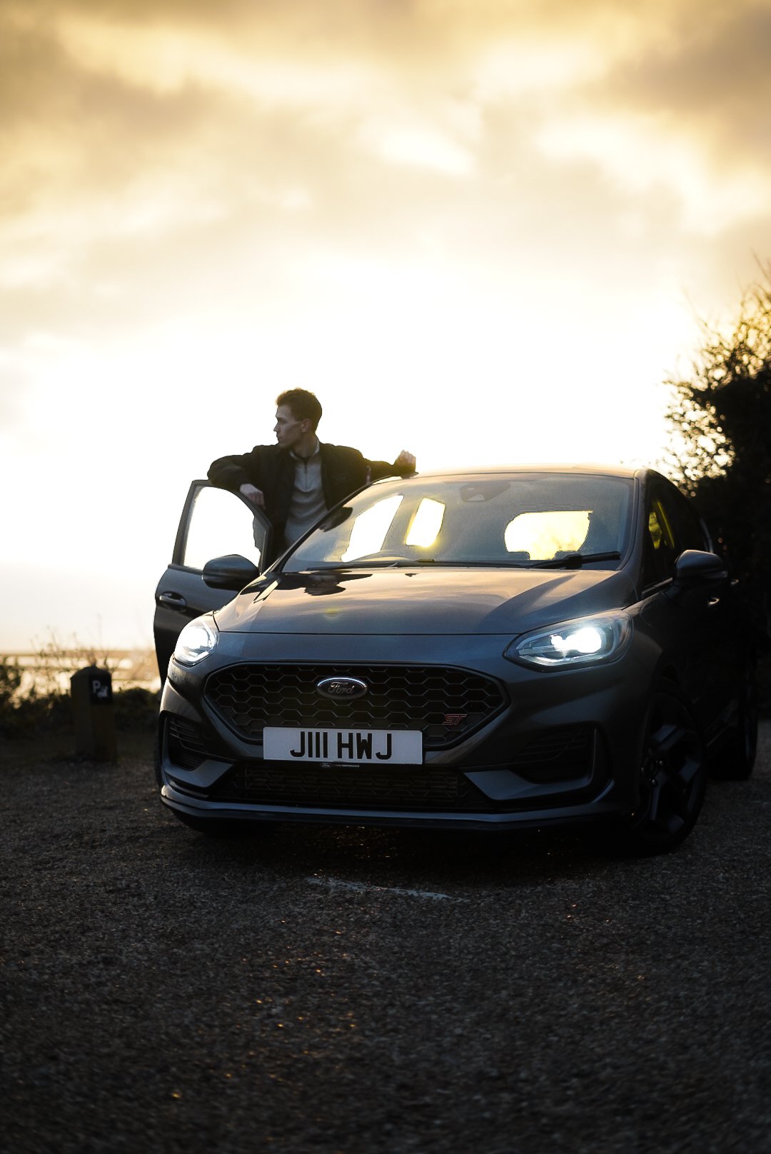 A man standing beside a black Ford car with the driver's door open, parked on a gravel surface during sunset or sunrise.