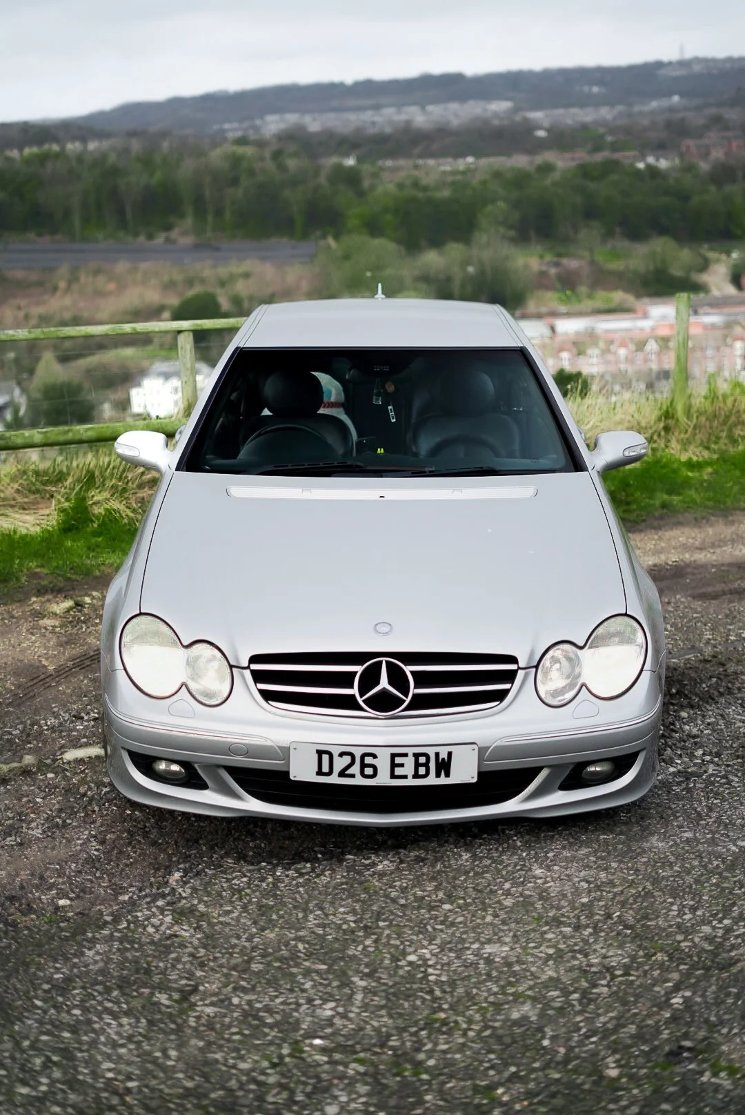 Front view of a silver Mercedes-Benz parked on a gravel surface with a green landscape and hills in the background.