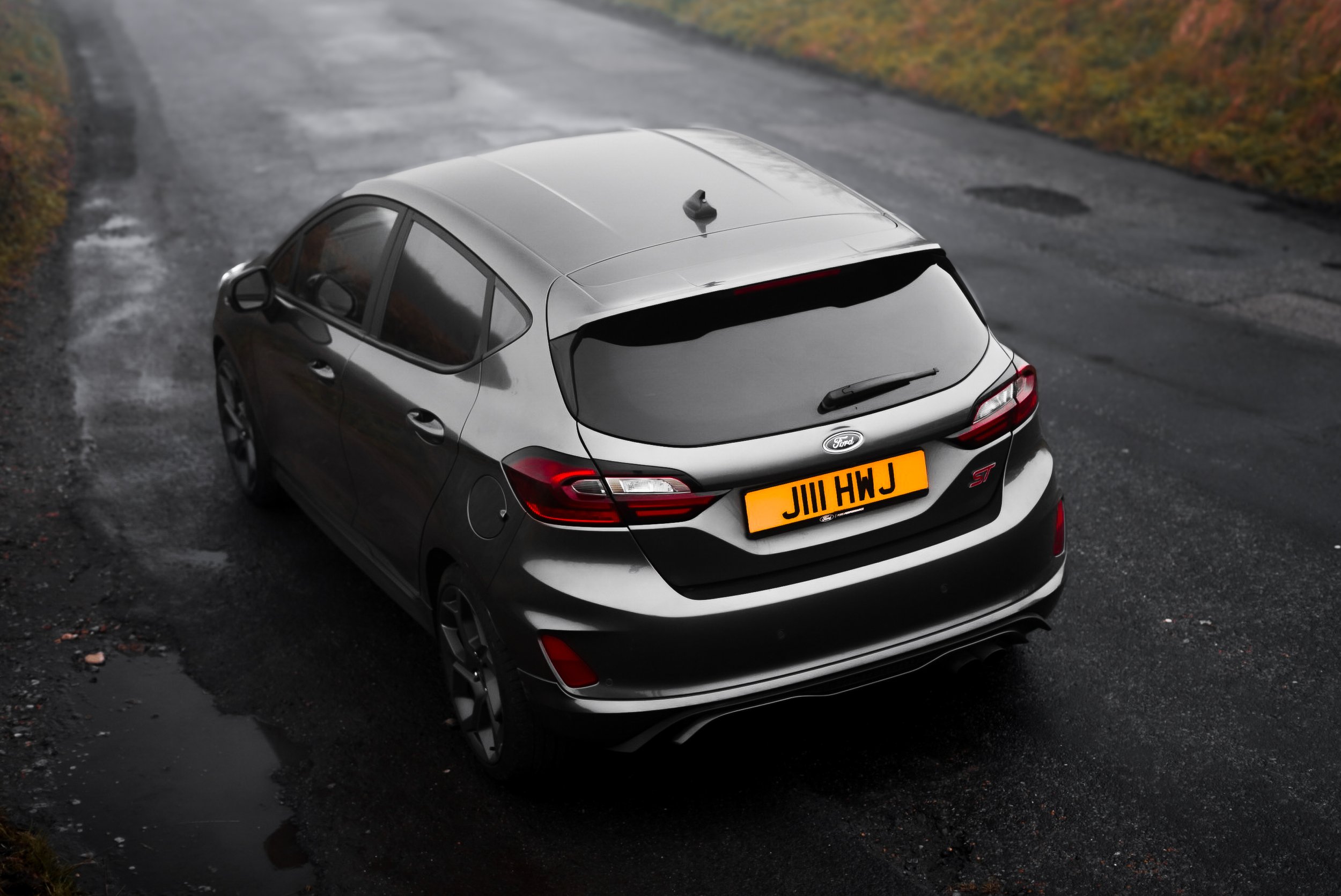 A silver Ford Fiesta ST hatchback parked on a wet, dark asphalt road with puddles, surrounded by fall foliage on the sides.