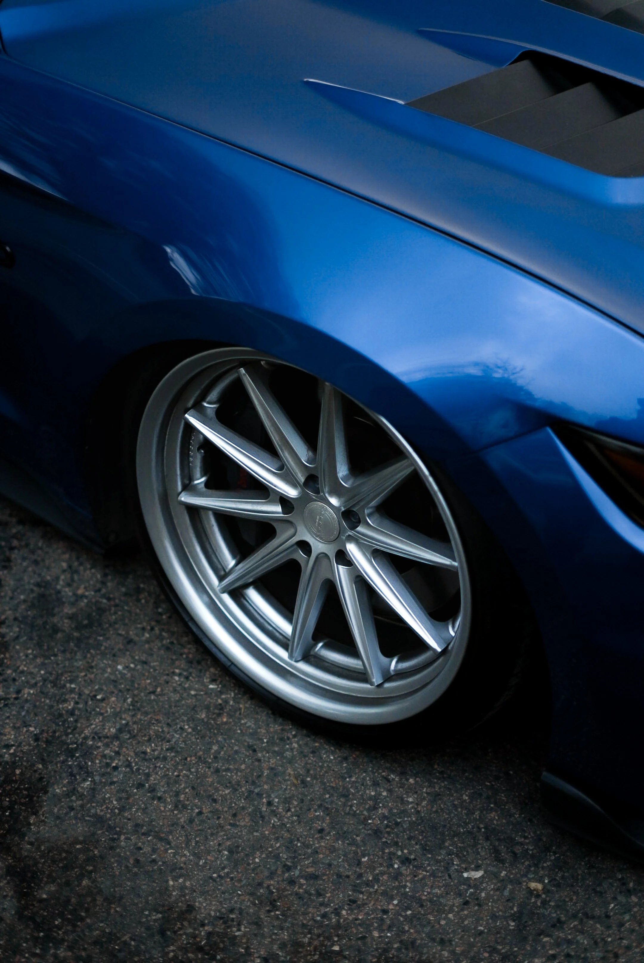 Close-up of a blue sports car with silver multi-spoke alloy wheels on a dark asphalt surface.