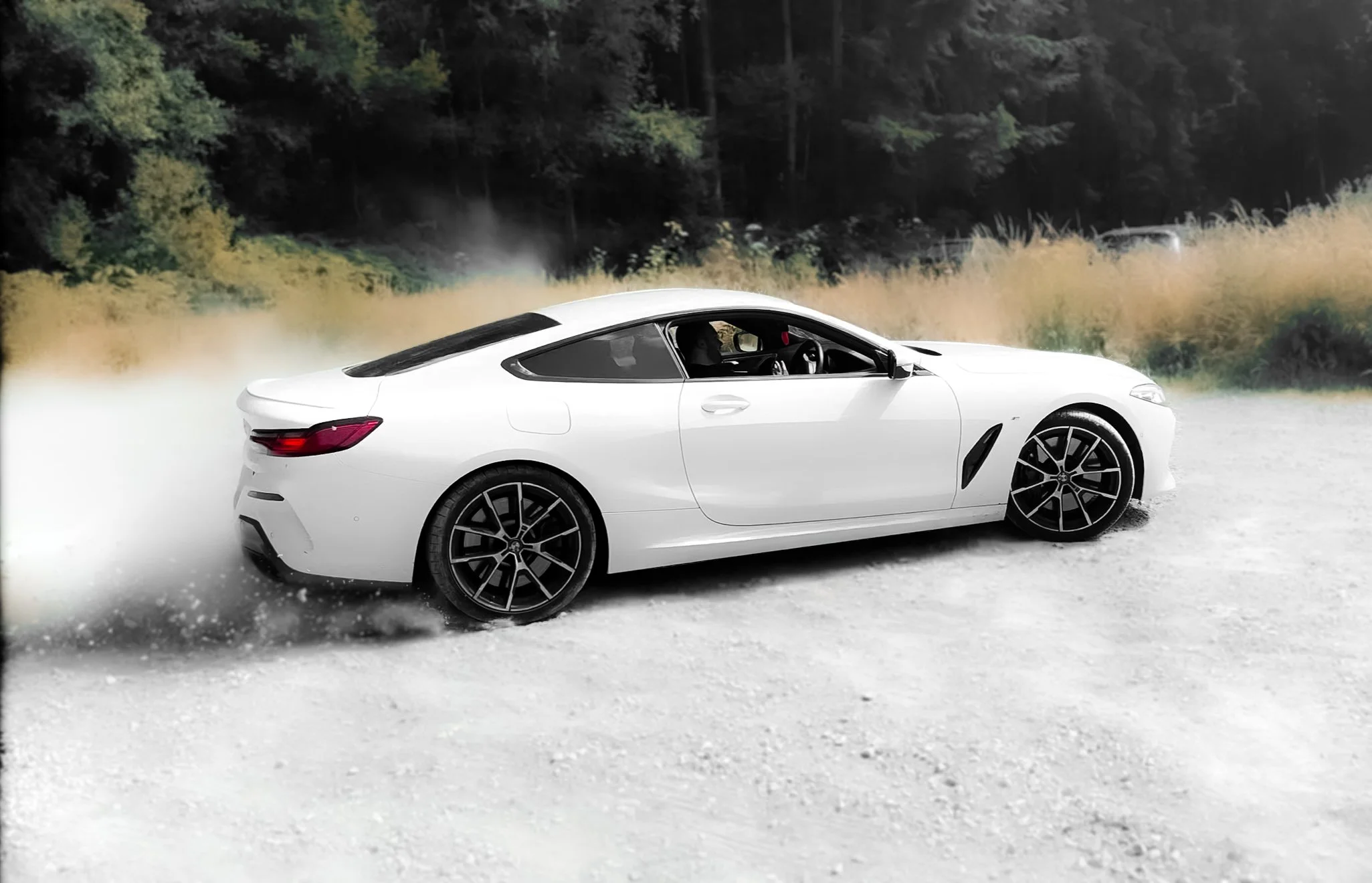 A white luxury sports car driving on a gravel road with dust and gravel kicking up behind it, surrounded by trees and grassy areas in the background.