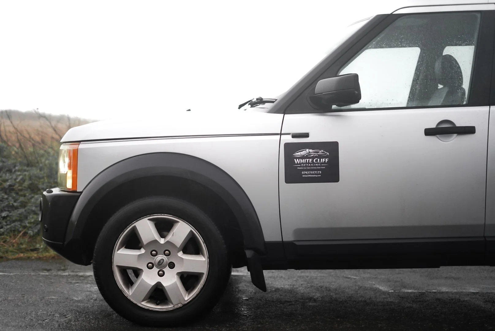 Silver vehicle with a black side mirror and black wheel arch. A black and white sticker on the door reads 'White Cliff Detailing' with a car logo, phone number, and website. The background is overcast and rainy, with sparse vegetation.