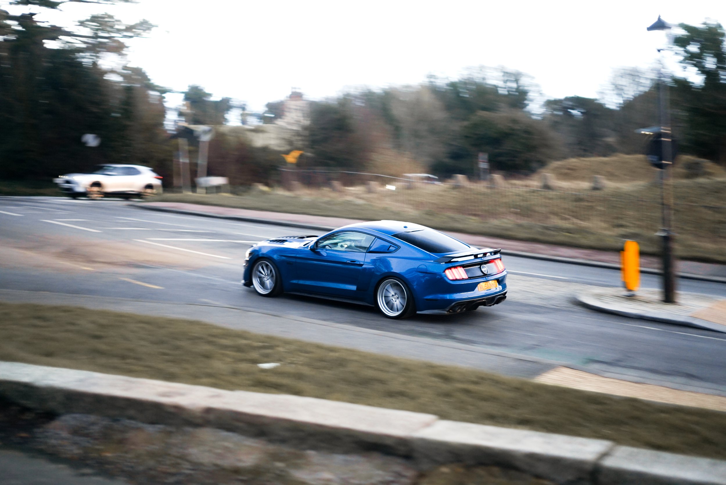 A blue sports car driving on a city street during daytime, with a blurred background of trees and other vehicles.